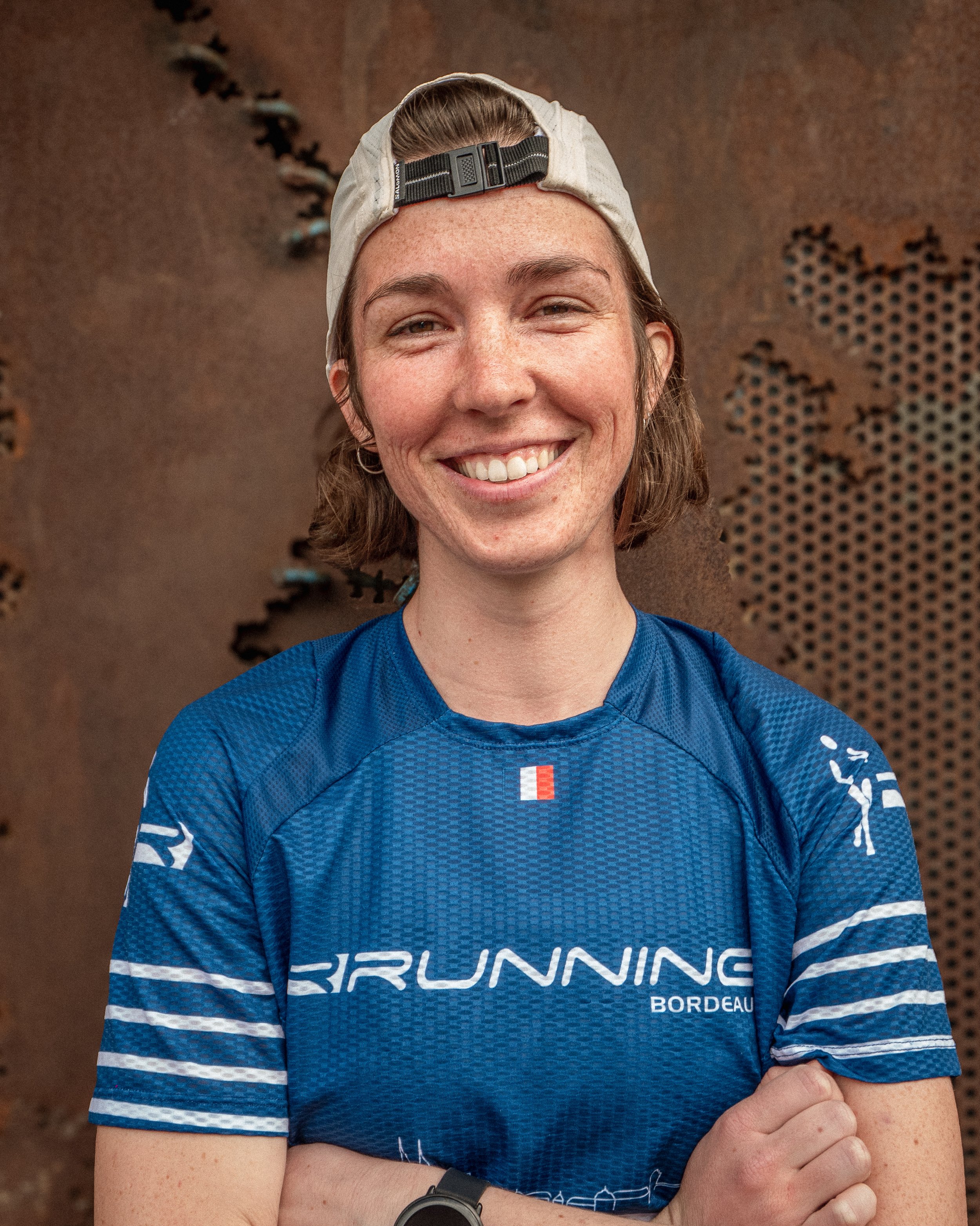 Jeune femme souriante portant une casquette beige à l'envers, une t-shirt de course bleue avec le mot 'Running Bordeaux', debout devant un mur en fer avec une carte du monde découpée.