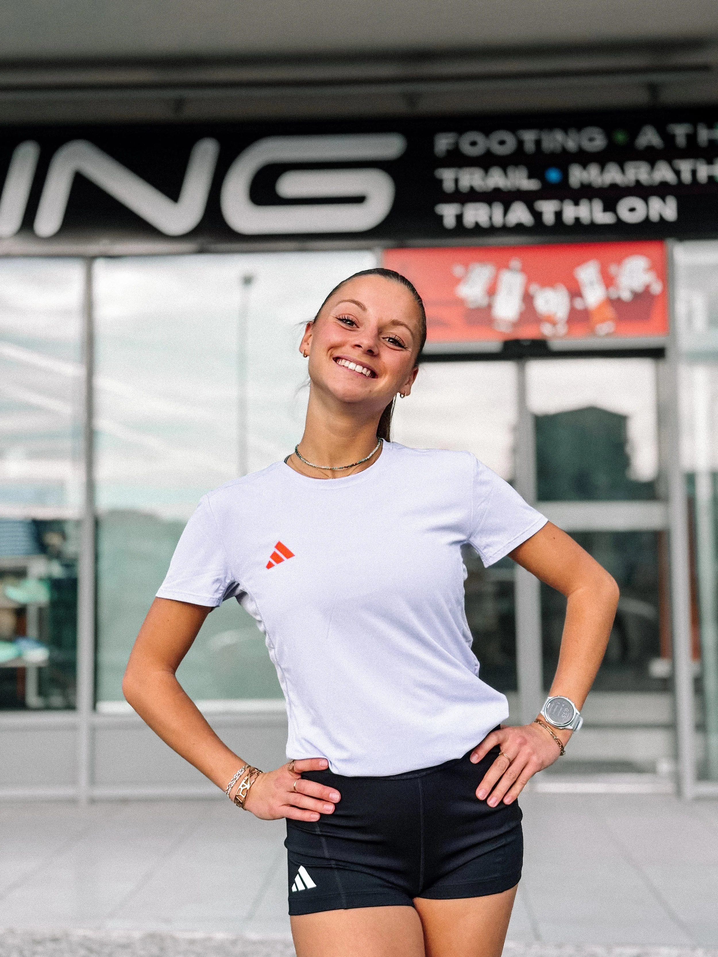 Jeune femme souriante en tenue de sport, debout devant un centre sportif, portant un T-shirt blanc, shorts noirs, montre et bracelets, avec un panneau indiquant différents sports comme course, trail, marathon et triathlon en arrière-plan.