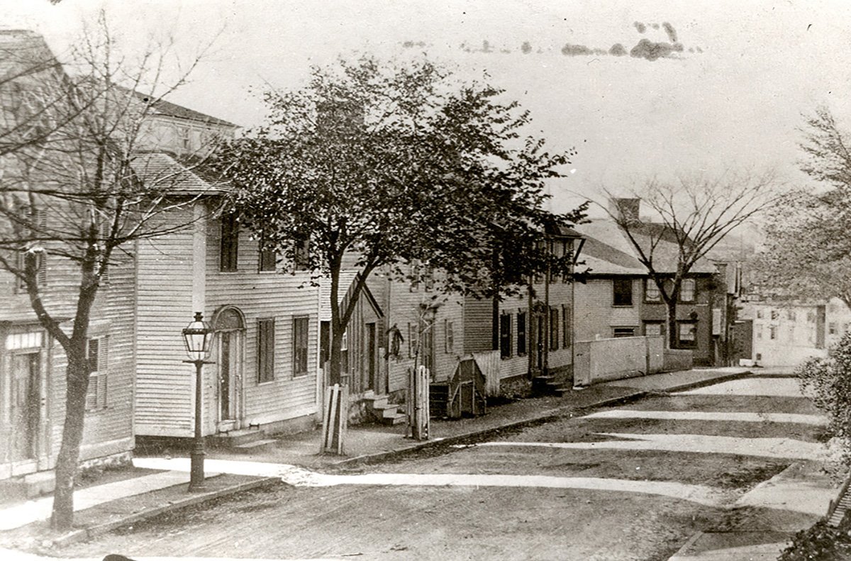 Black and white photo of a residential street with old-style wooden houses, leafless and leafy trees, street lamps, and a dirt road with curving tracks.