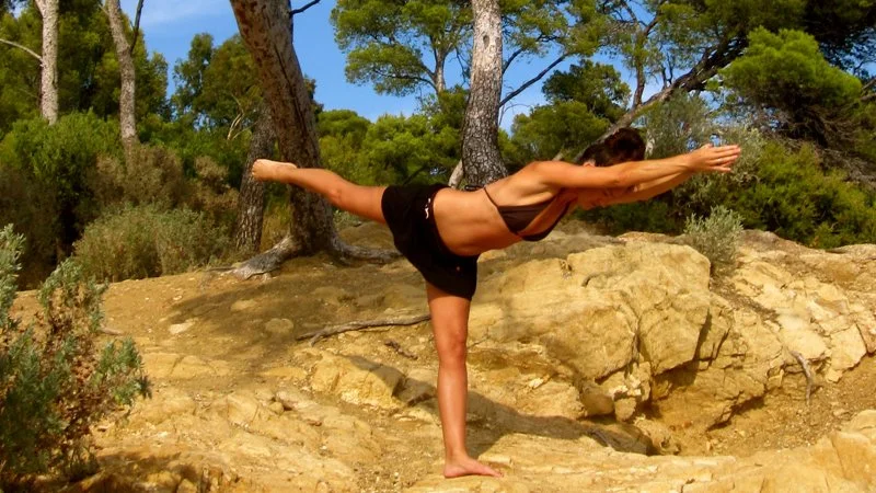 Une femme pratique le yoga en équilibre sur une jambe dans un environnement naturel avec des arbres et des rochers.