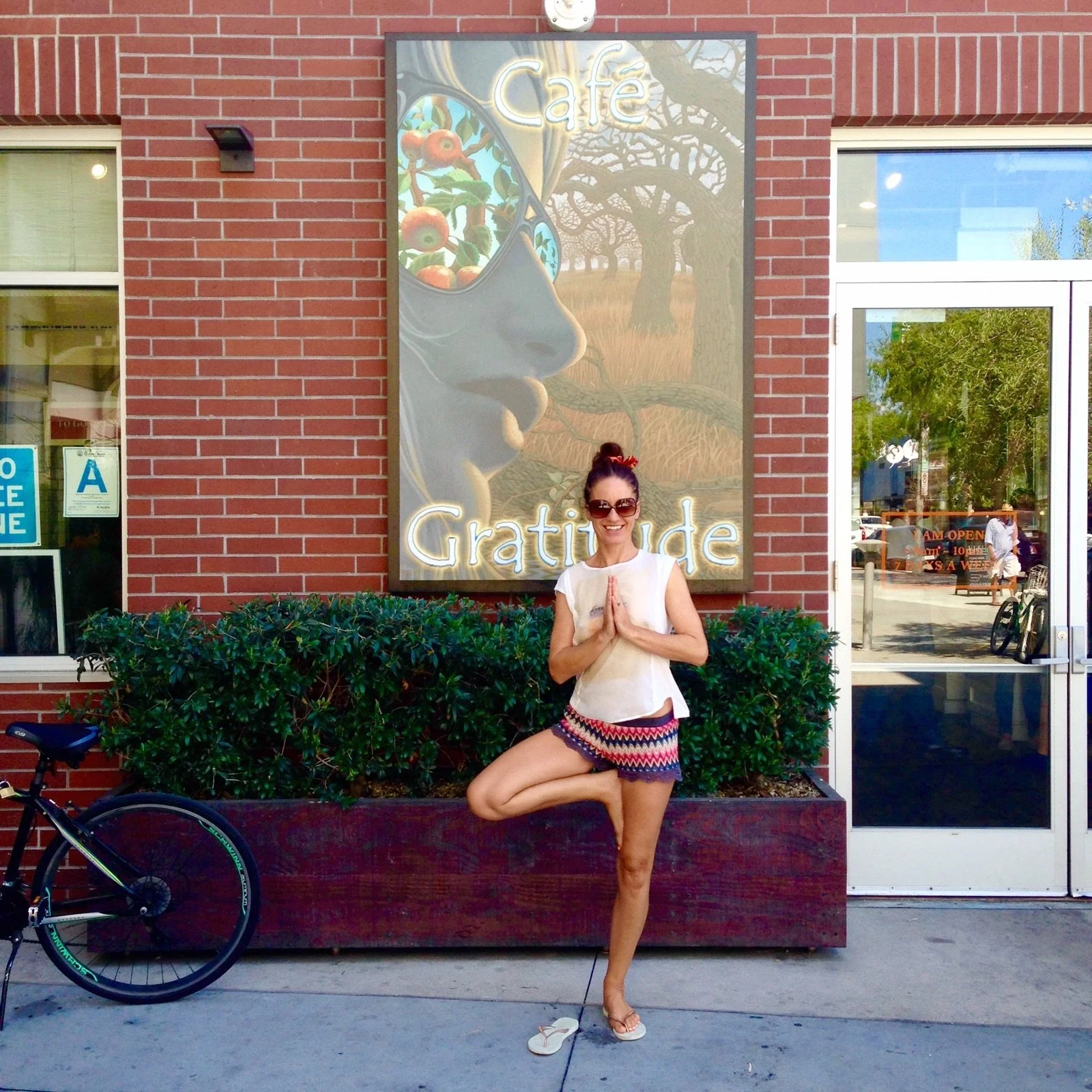 Une femme en posture de yoga de l'arbre devant une enseigne de café avec le texte 'Café Gratitude', souriant et portant des lunettes de soleil, en été.