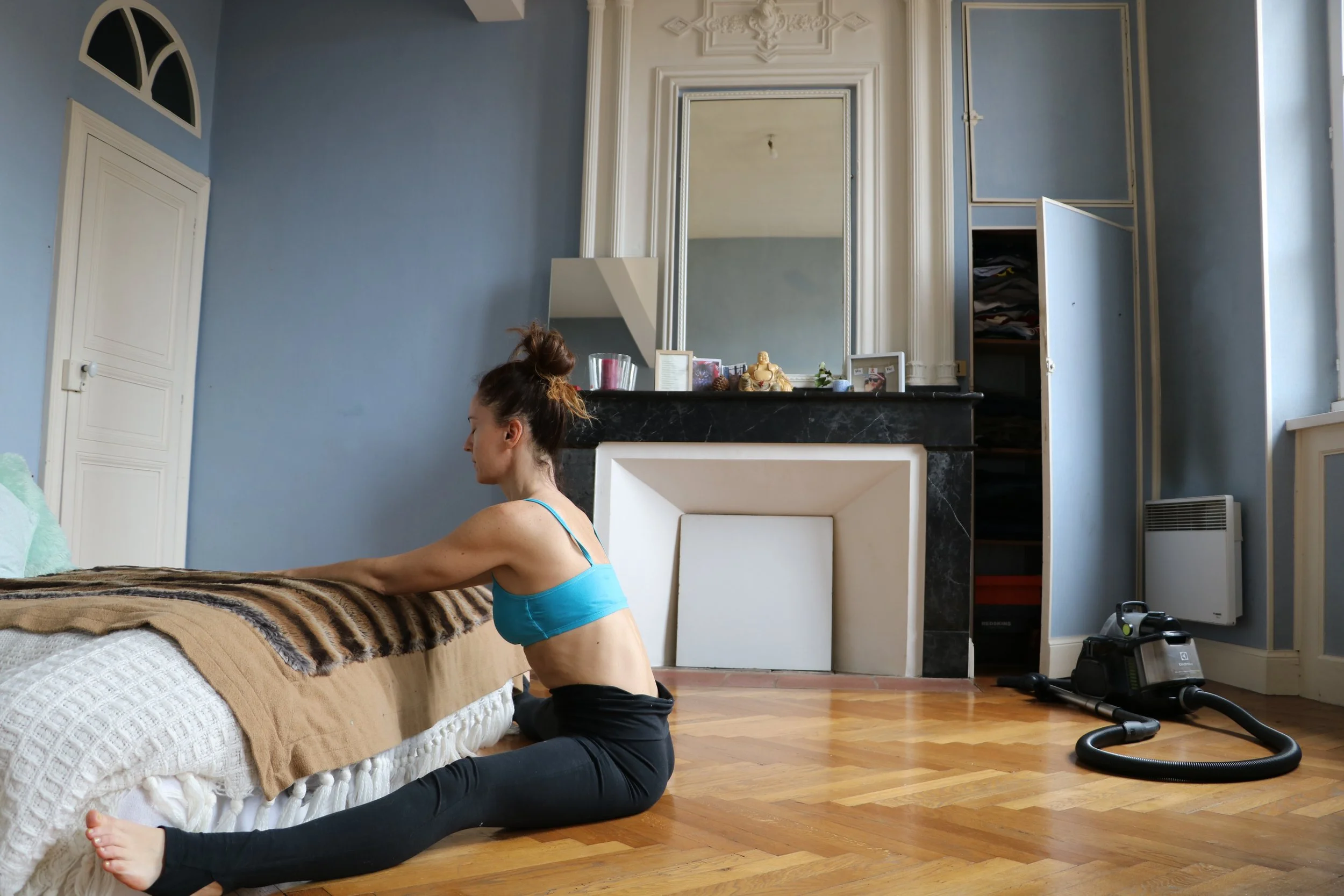 Une femme en vêtements de sport s'étire sur le sol dans un salon avec un parquet en bois, près d'un lit. Le salon a un mur bleu, une cheminée noire avec un grand miroir au-dessus, des étagères ouvertes, un radiateur, et un aspirateur sur le sol.