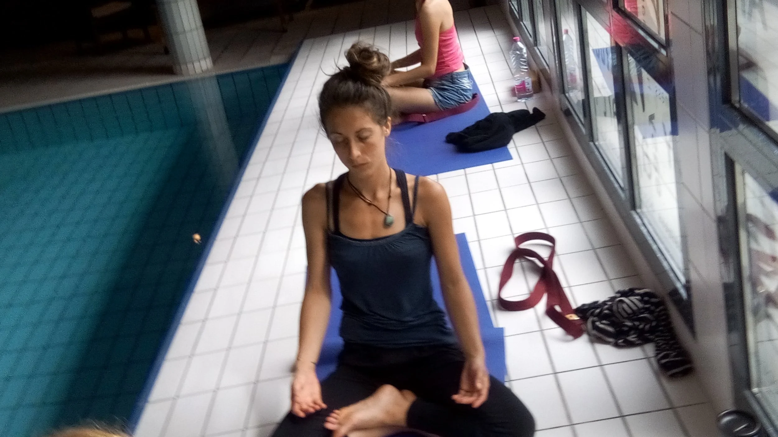 Deux femmes pratiquent le yoga près d'une piscine, assises sur des tapis avec des objets personnels à côté, dans un espace intérieur avec grandes fenêtres.