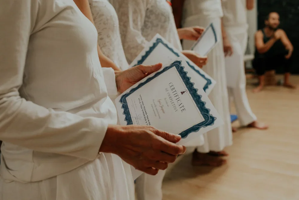 Groupe de yoginis habillées en blanc en train de recevoir des certificats de professeur de yoga lors d'une cérémonie de clôture de formation de yoga. Un homme en arrière-plan est assis, habillé en noir.