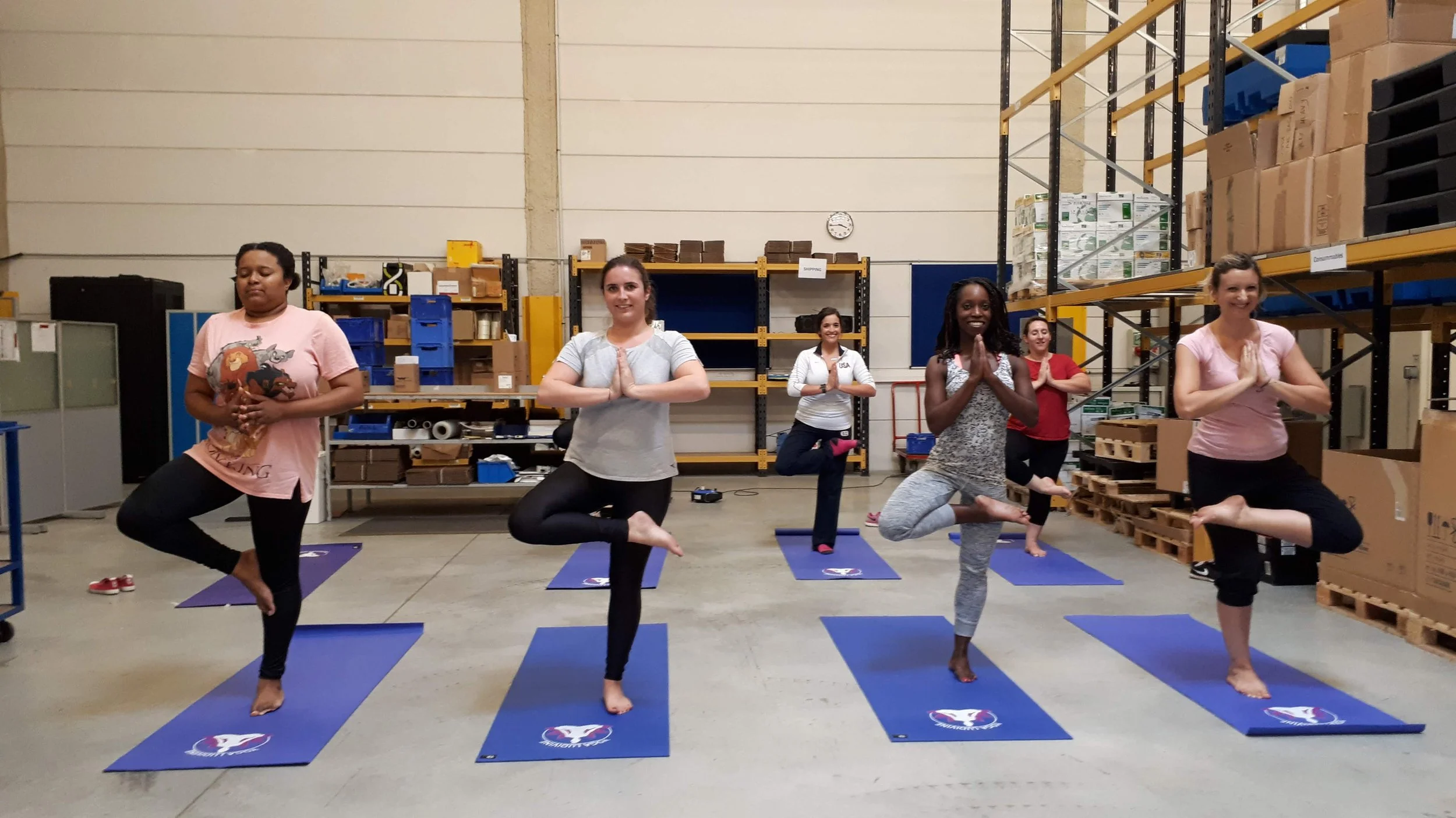 Groupe de femmes faisant du yoga en entreprise dans une salle de stockage ou d'entrepôt. Elles sont en position d'arbre, sur leurs tapis de yoga.
