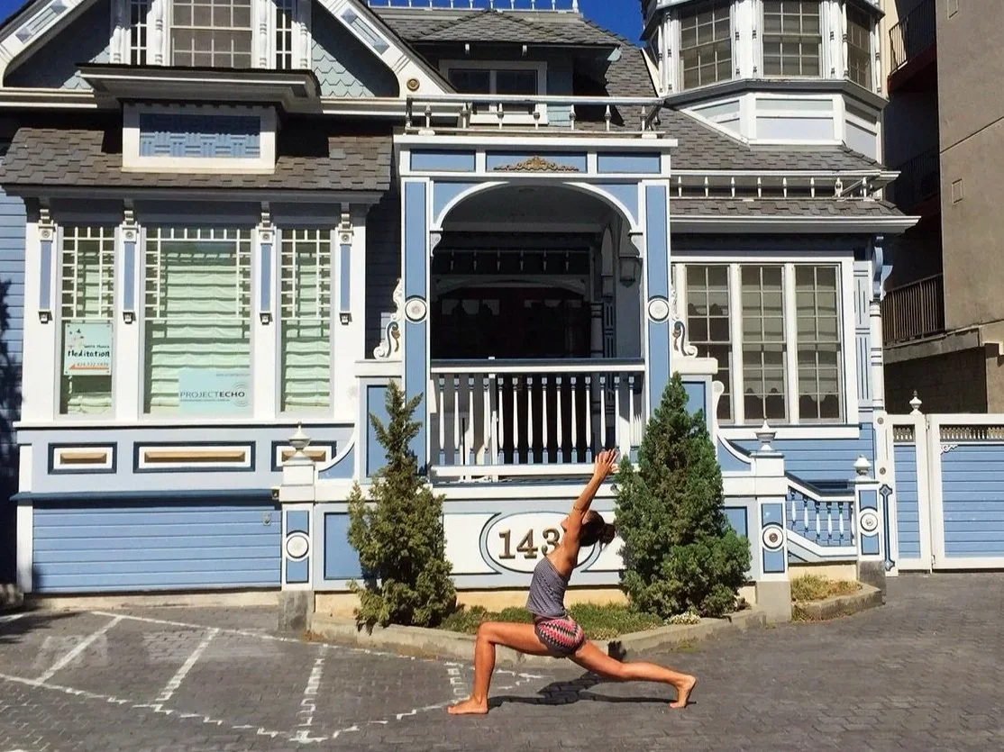 Femme en position de yoga dans une rue devant une grande maison victorienne bleue avec des détails blancs, devant un jardin avec deux petits arbres et un numéro 143. En Californie.