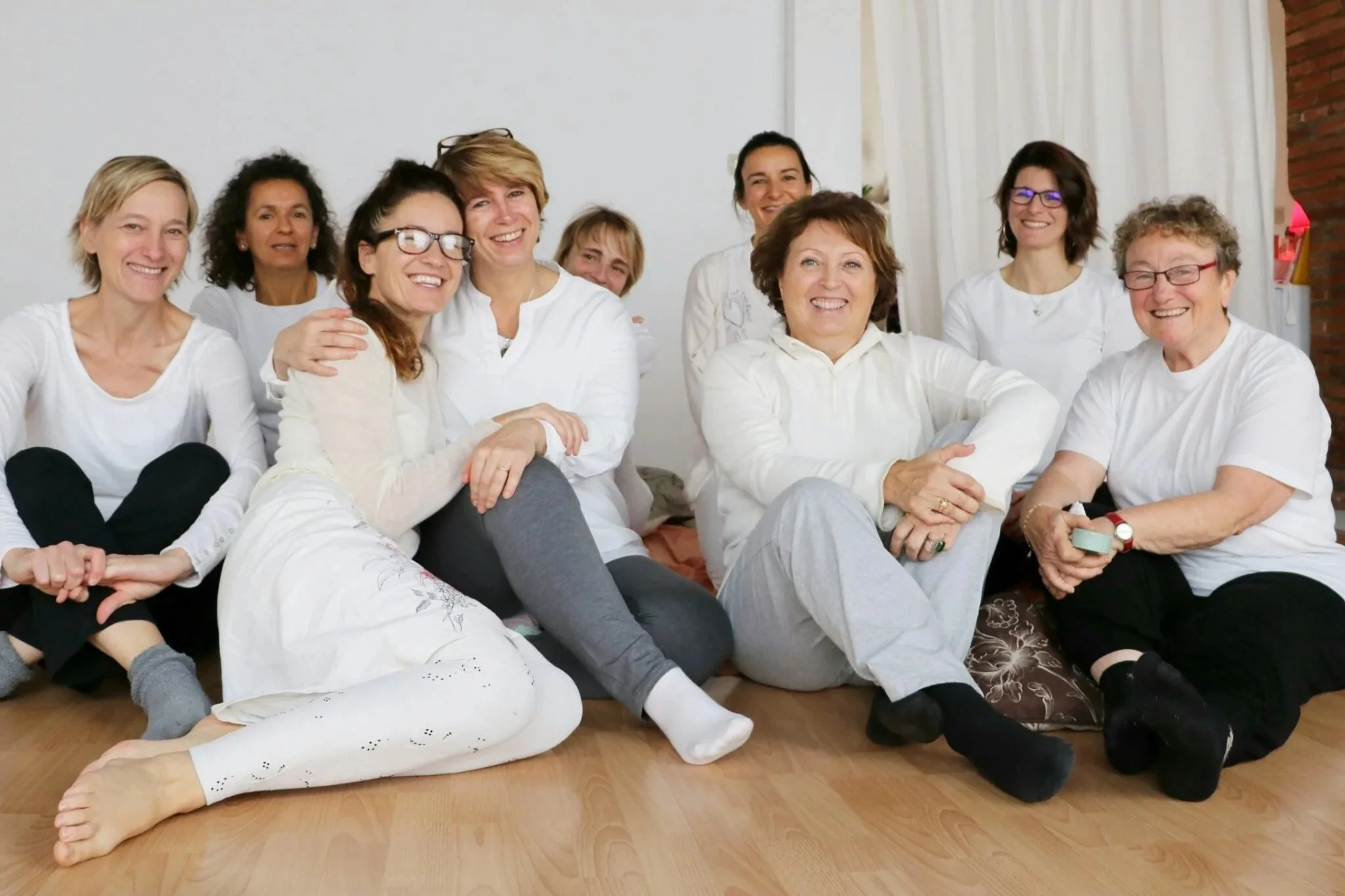 Groupe de femmes assises par terre, souriantes, dans une pièce lumineuse avec un mur blanc et un rideau blanc derrière elles.