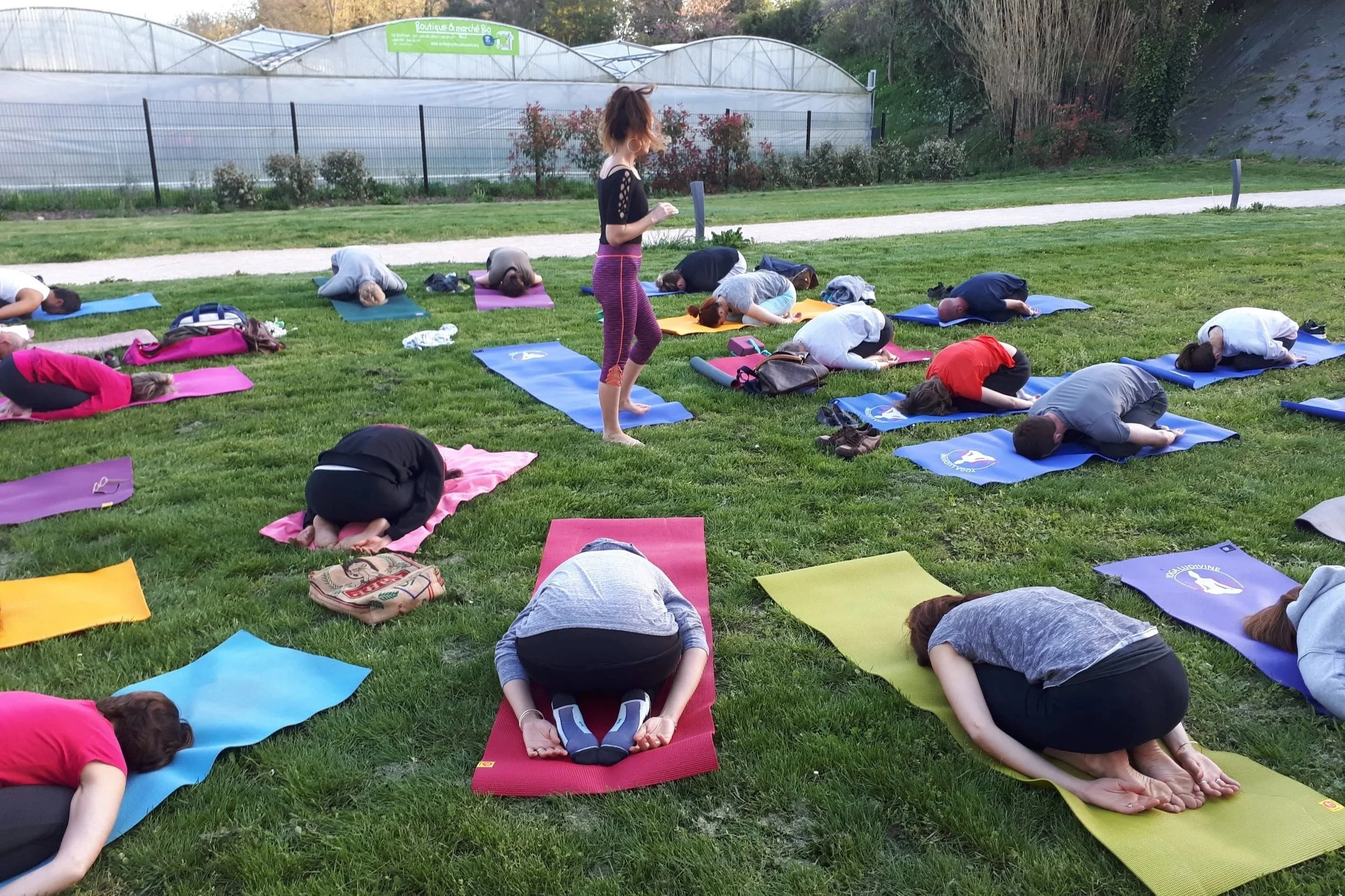 Groupe de personnes participant à une séance de yoga en extérieur, pratiquant la position du Child's Pose sur des tapis colorés dans un espace vert, avec un instructeur debout au centre.