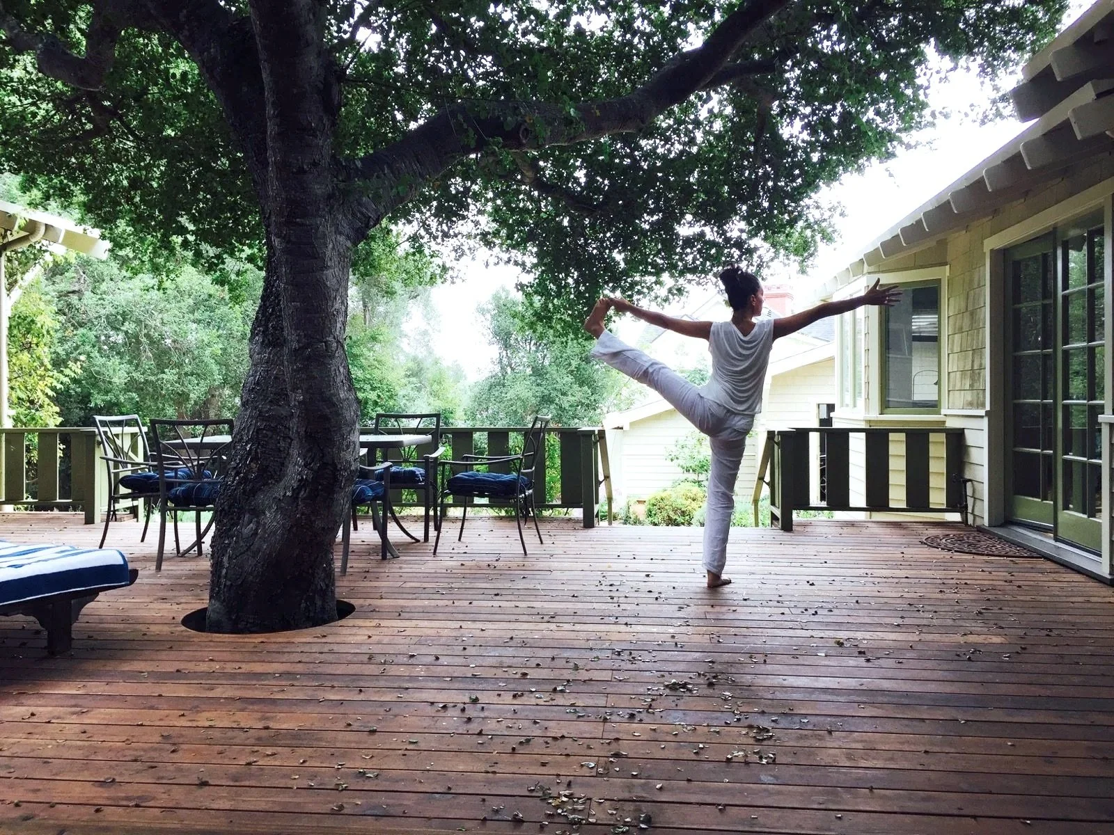 Une femme pratique le yoga en équilibre sur une jambe sur une terrasse en bois, entourée d'arbres et de mobilier d'extérieur.