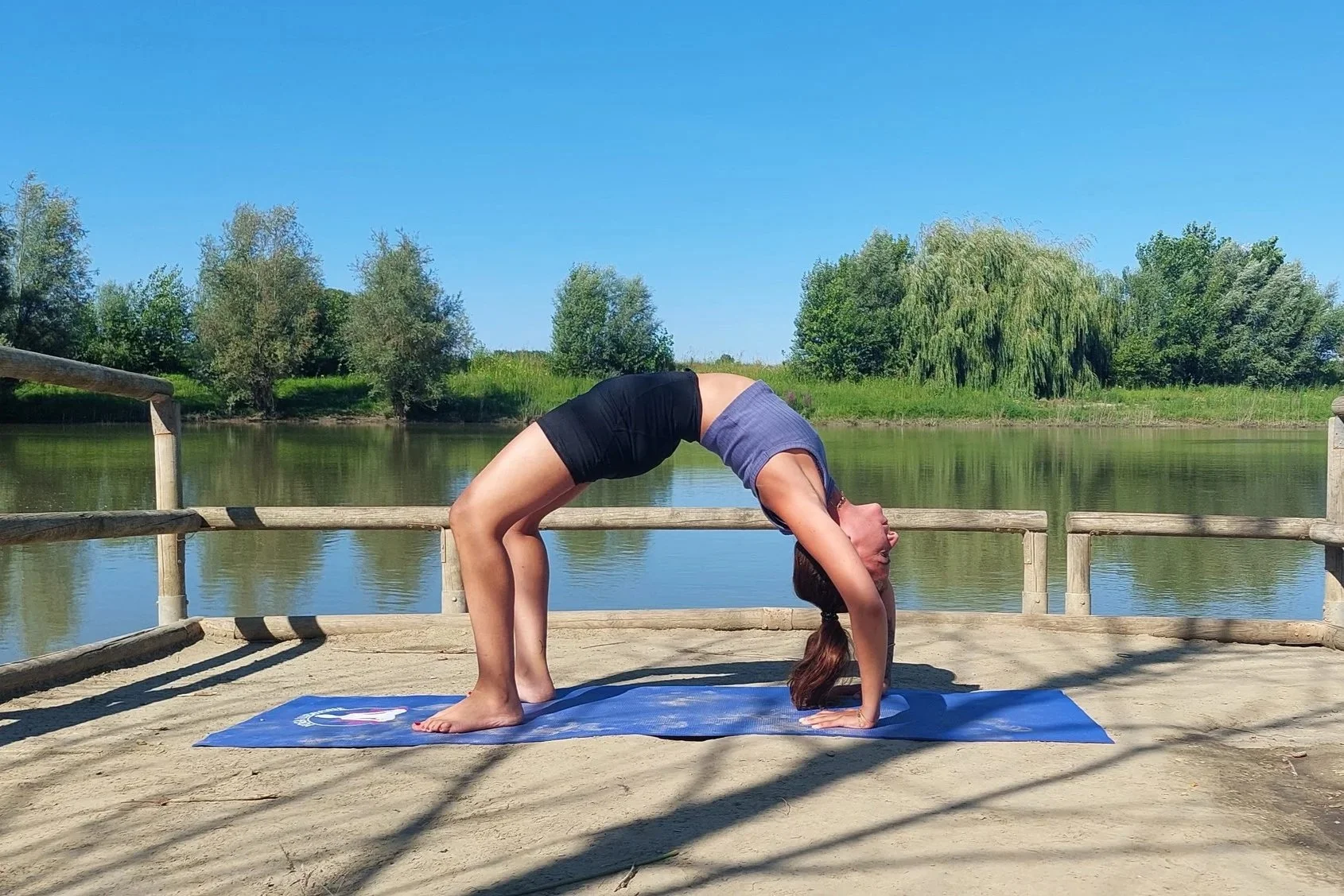 Femme faisant la posture du pont en yoga en plein air sur un pont en bois au bord de l'eau, avec des arbres en arrière-plan et un ciel bleu.