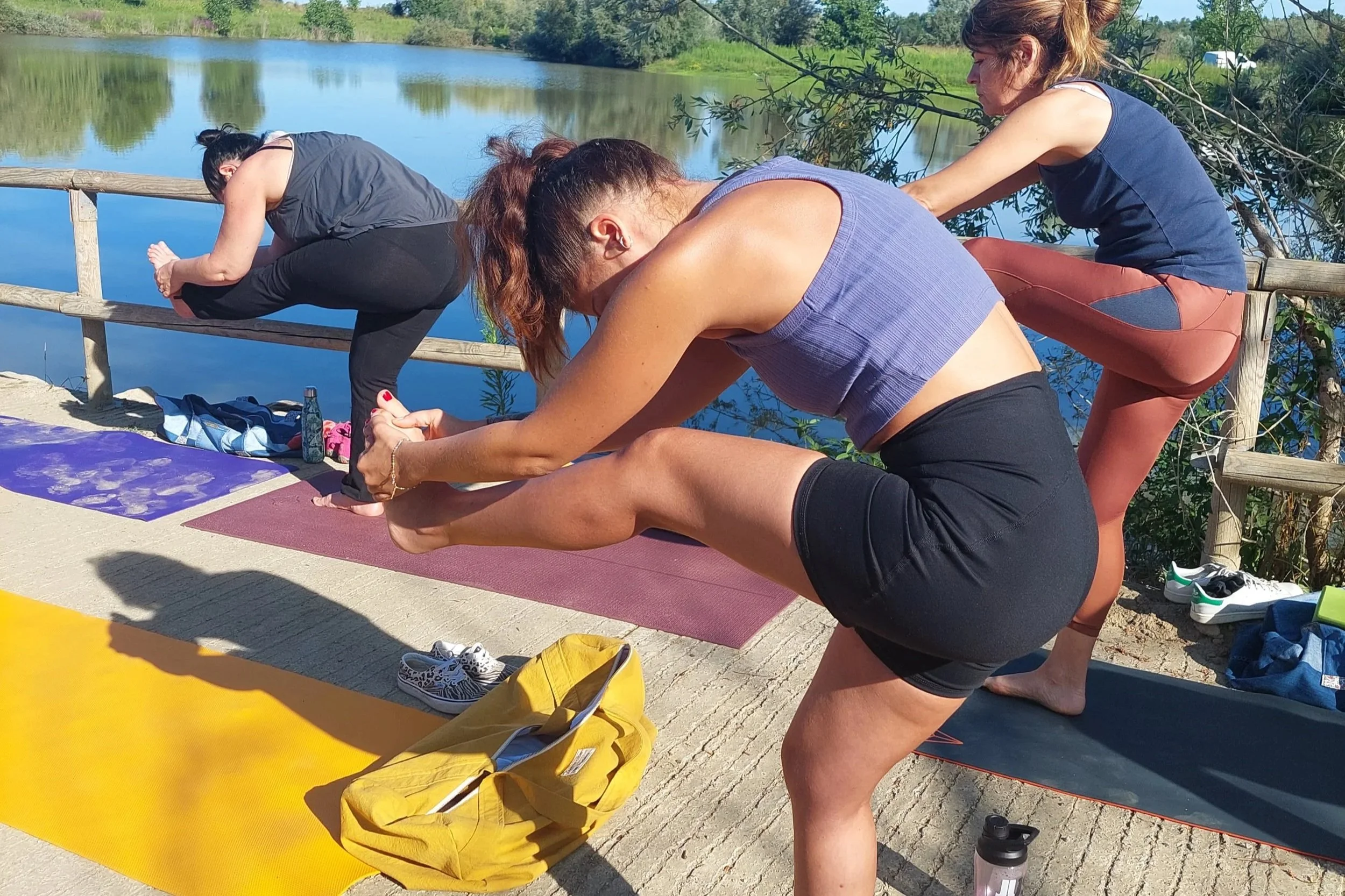 Trois femmes faisant du yoga en plein air près d'un lac en position de posture de flexion avant, avec des tapis de yoga sur le sol, un sac à dos jaune, des chaussures et des bouteilles d'eau à proximité.