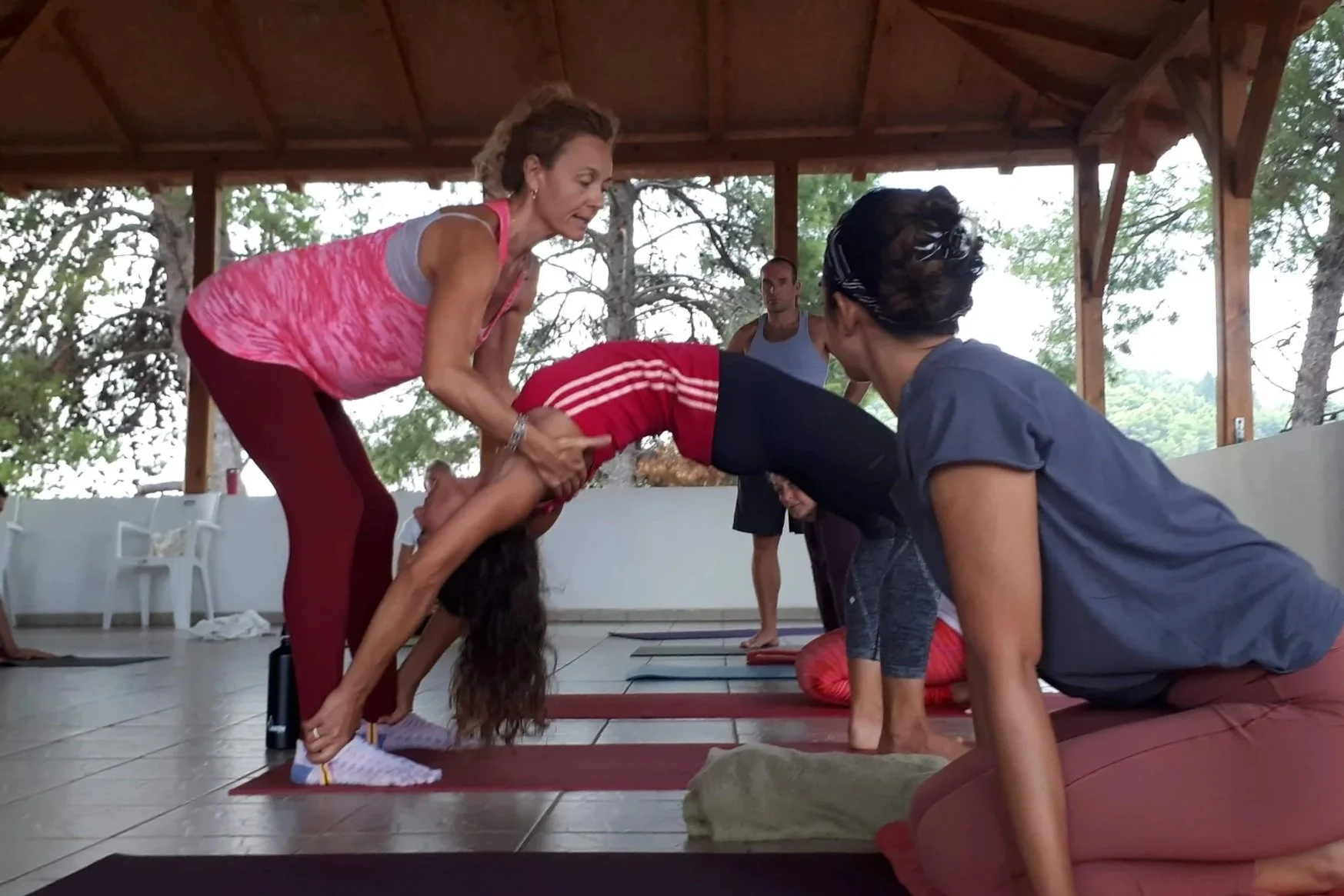 Groupe de personnes faisant du yoga en position de ponte, aidé par deux instructrices dans un espace couvert avec vue sur la nature.