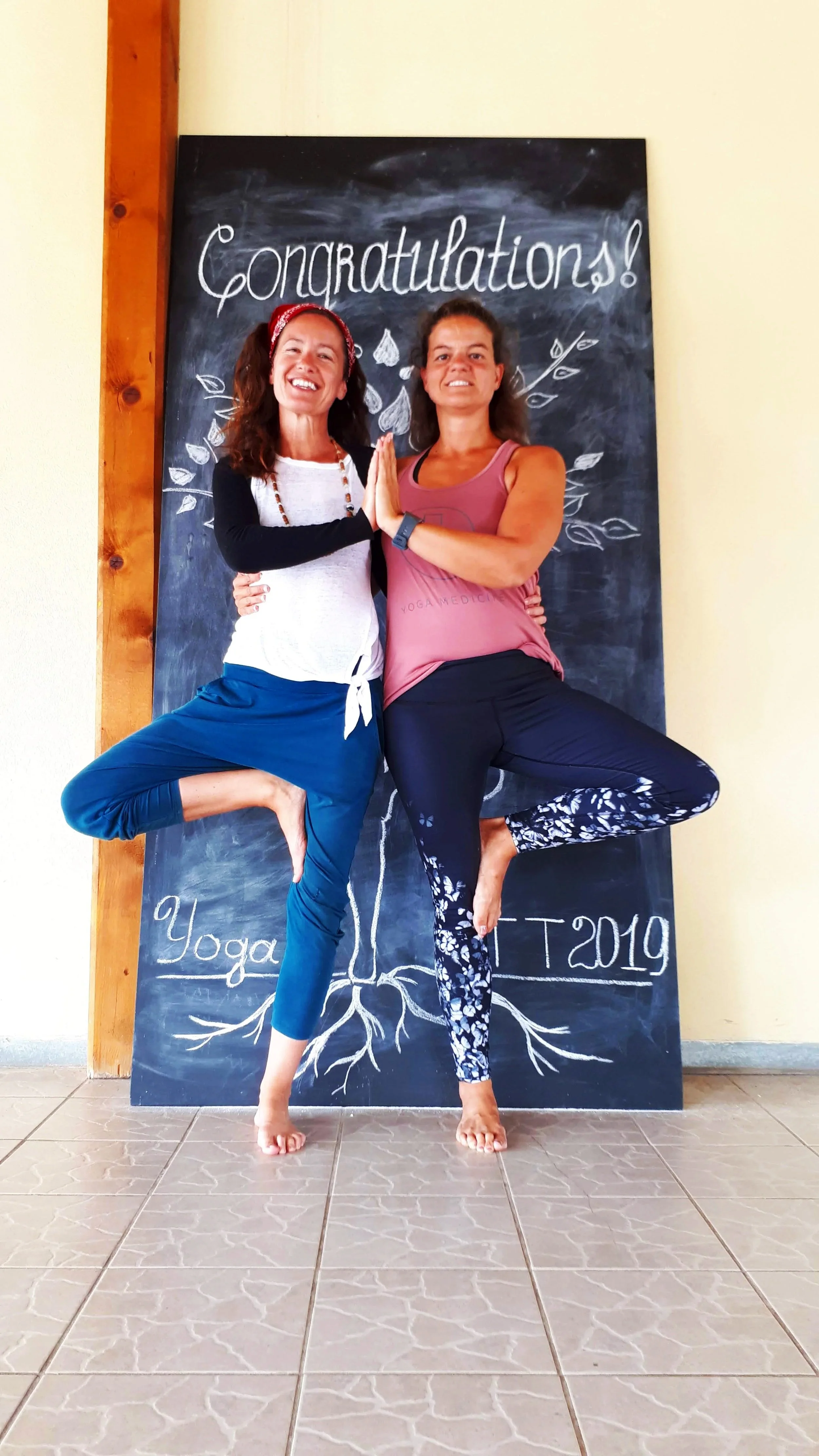 Deux femmes pratiquant le yoga en équilibre sur une jambe, souriantes, en intérieur devant un tableau noir avec des écritures et dessins de feuilles et un arbre. L'une porte un t-shirt blanc et un pantalon bleu, l'autre porte un débardeur rose et un 