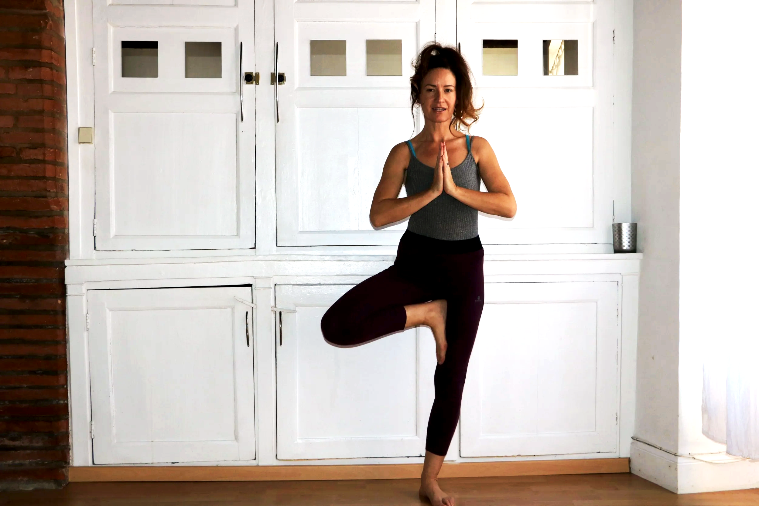 Femme pratiquant la posture de l'arbre en yoga dans une pièce avec un mur blanc et une armoire en bois.