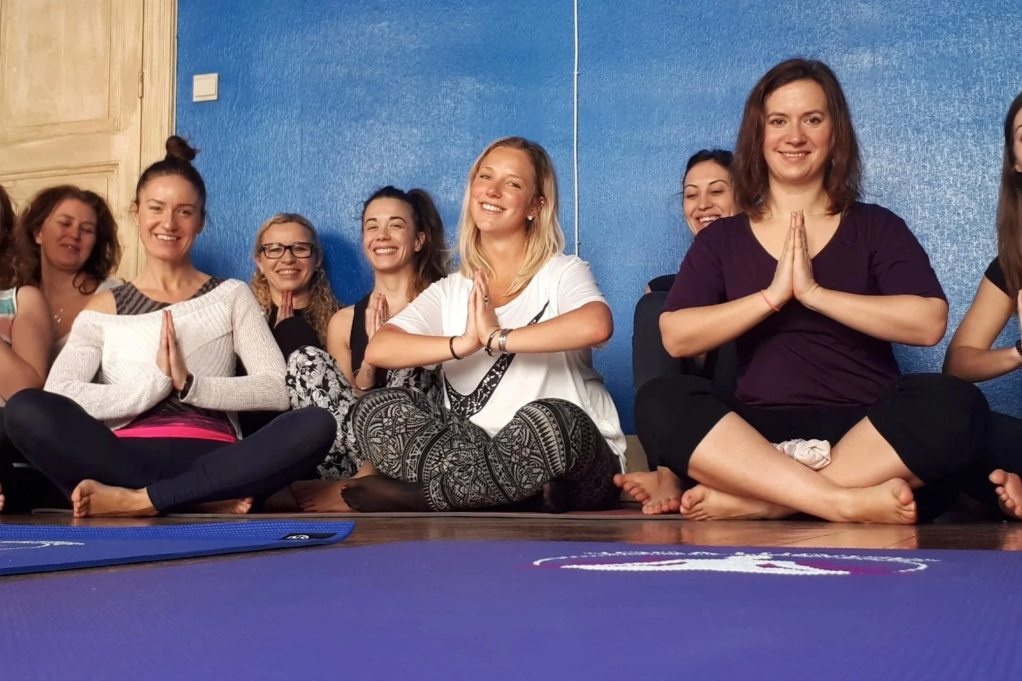 Groupe de femmes assises par terre, pratiquant la méditation ou le yoga, avec les mains en position de prière devant la poitrine, dans une pièce avec un mur bleu.