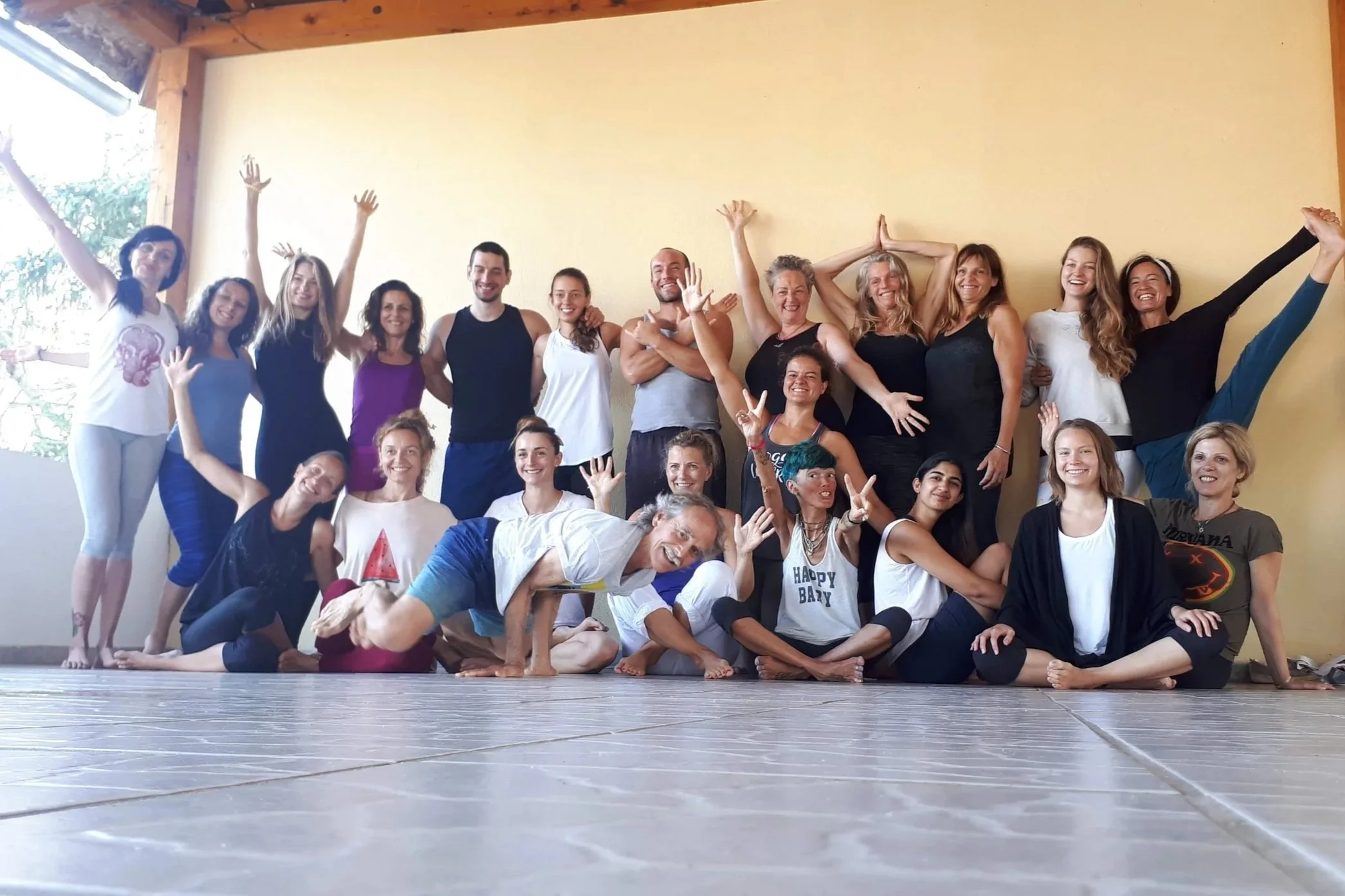 Groupe de personnes souriantes posant dans une salle avec un mur beige et des poutres en bois, certains en position assise ou allongée, d'autres en train de se tenir debout, en diverses poses joyeuses.