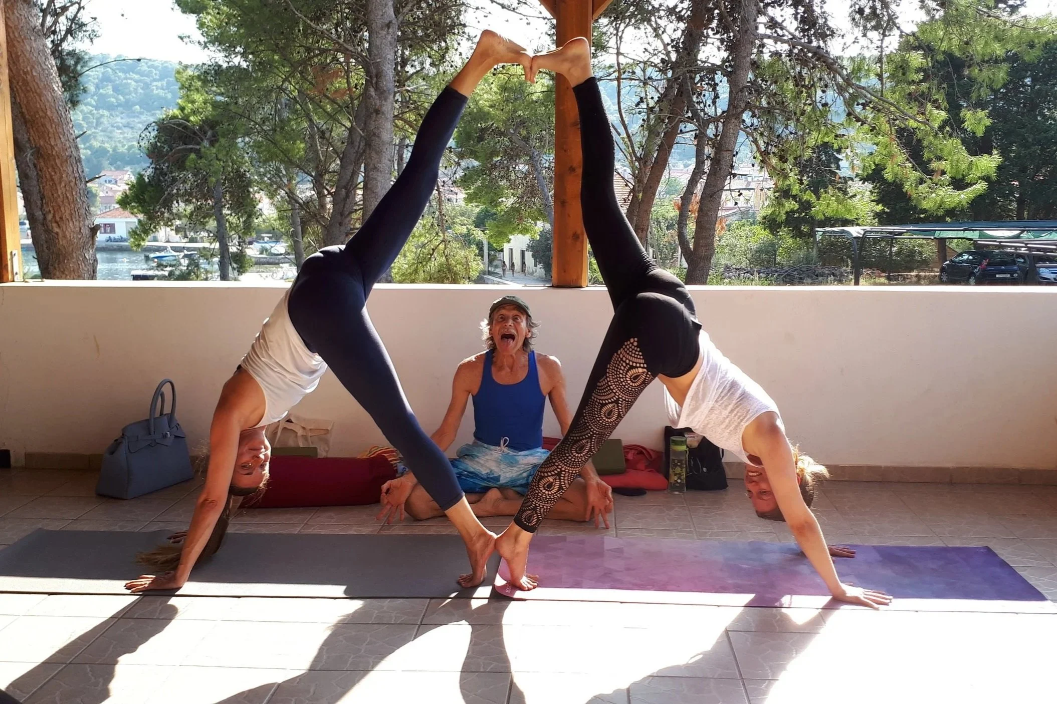 Deux femmes faisant du yoga en position de roue inversée avec un instructeur assis au centre, dans un espace couvert avec vue sur la nature et des arbres en arrière-plan.