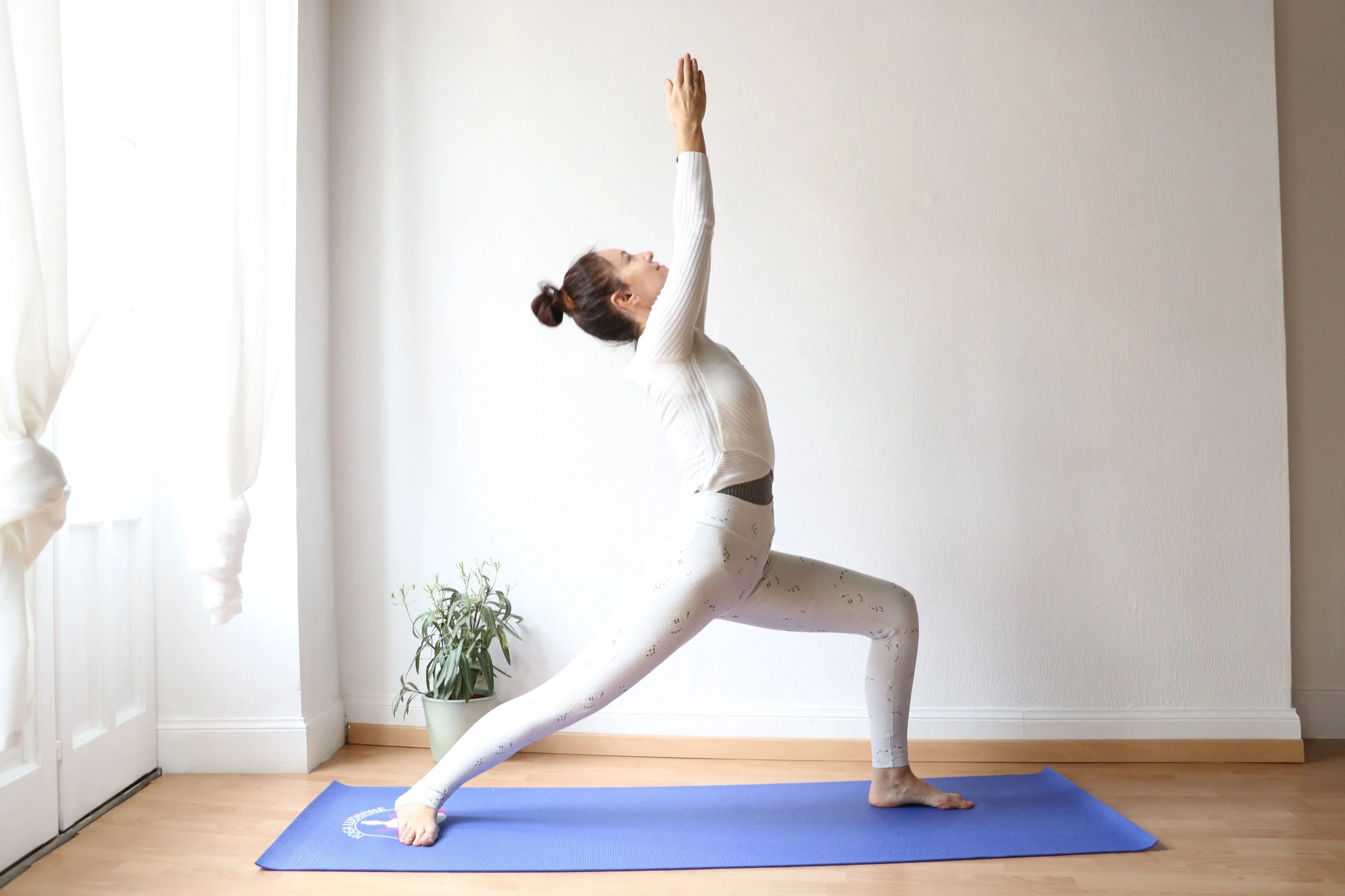 Femme faisant du yoga en position guerrier sur un tapis bleu dans une pièce lumineuse avec des rideaux blancs, une plante en pot à côté.