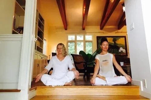 Deux femmes en méditation en position de lotus dans une maison lumineuse, assises sur un plancher en bois, avec des meubles et une peinture en arrière-plan.