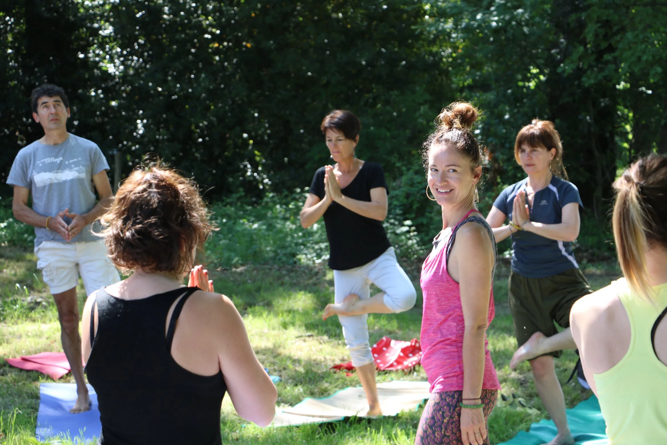 Groupe de personnes pratiquant le yoga en plein air dans un parc avec des arbres en arrière-plan, certaines en position de méditation ou de pose de yoga.