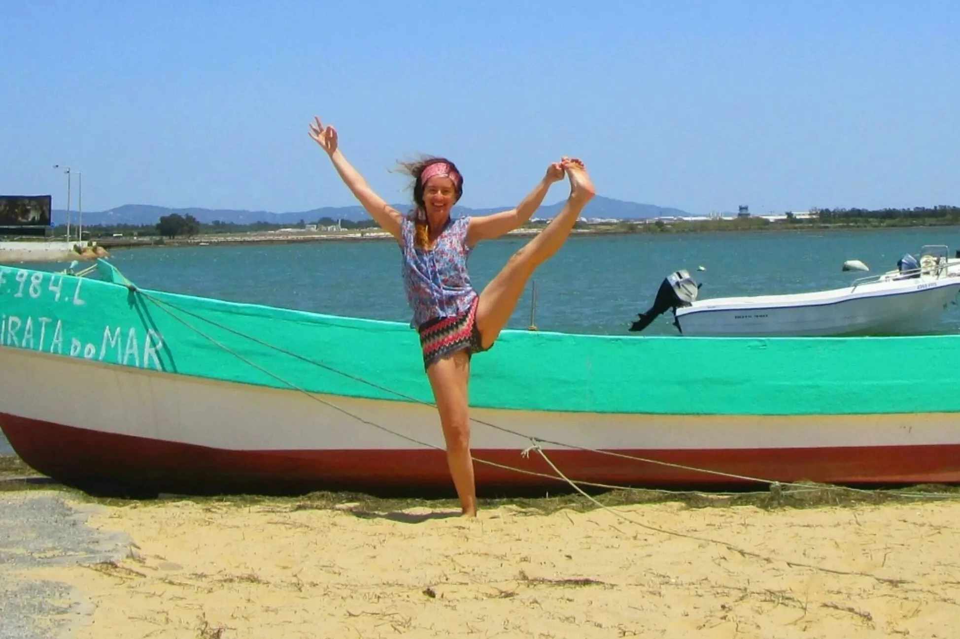 Une femme souriante en vêtements colorés, en yoga danse, debout sur une jambe en équilibre sur la plage, avec un bateau en arrière-plan sous un ciel ensoleillé.