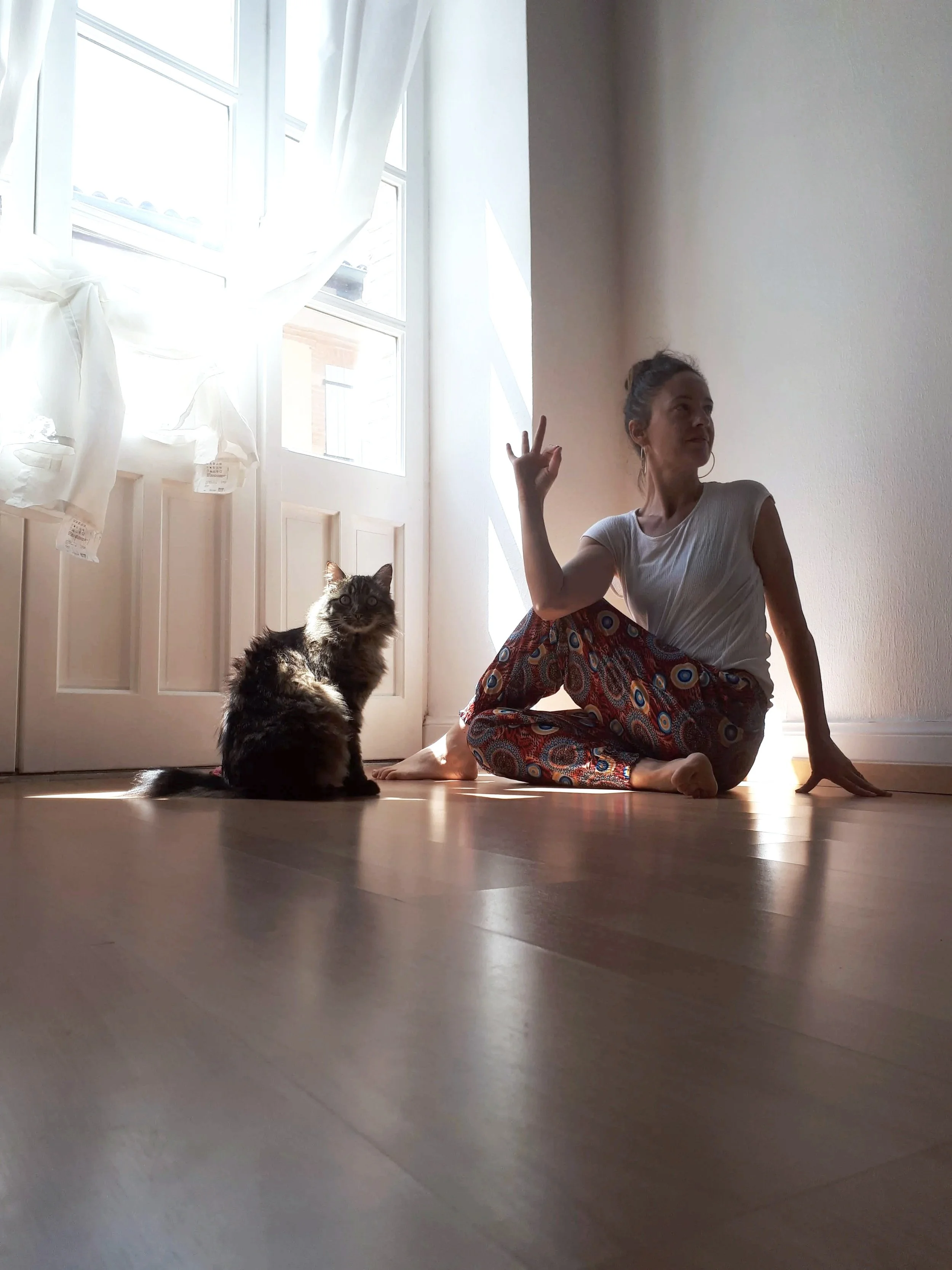 Une femme assise sur le sol en pyjama avec un motif coloré, faisant un geste avec la main, à côté d'un chat tacheté noir et gris, dans une pièce lumineuse avec une grande fenêtre et des rideaux blancs.