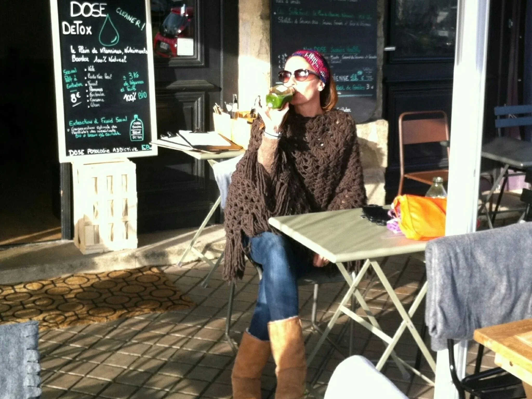 Ludivine assise à une table en terrasse à Bordeaux, portant un pull marron en crochet, des bottes en daim, des lunettes de soleil, et un bandeau rouge à motifs, en train de boire un jus vert.