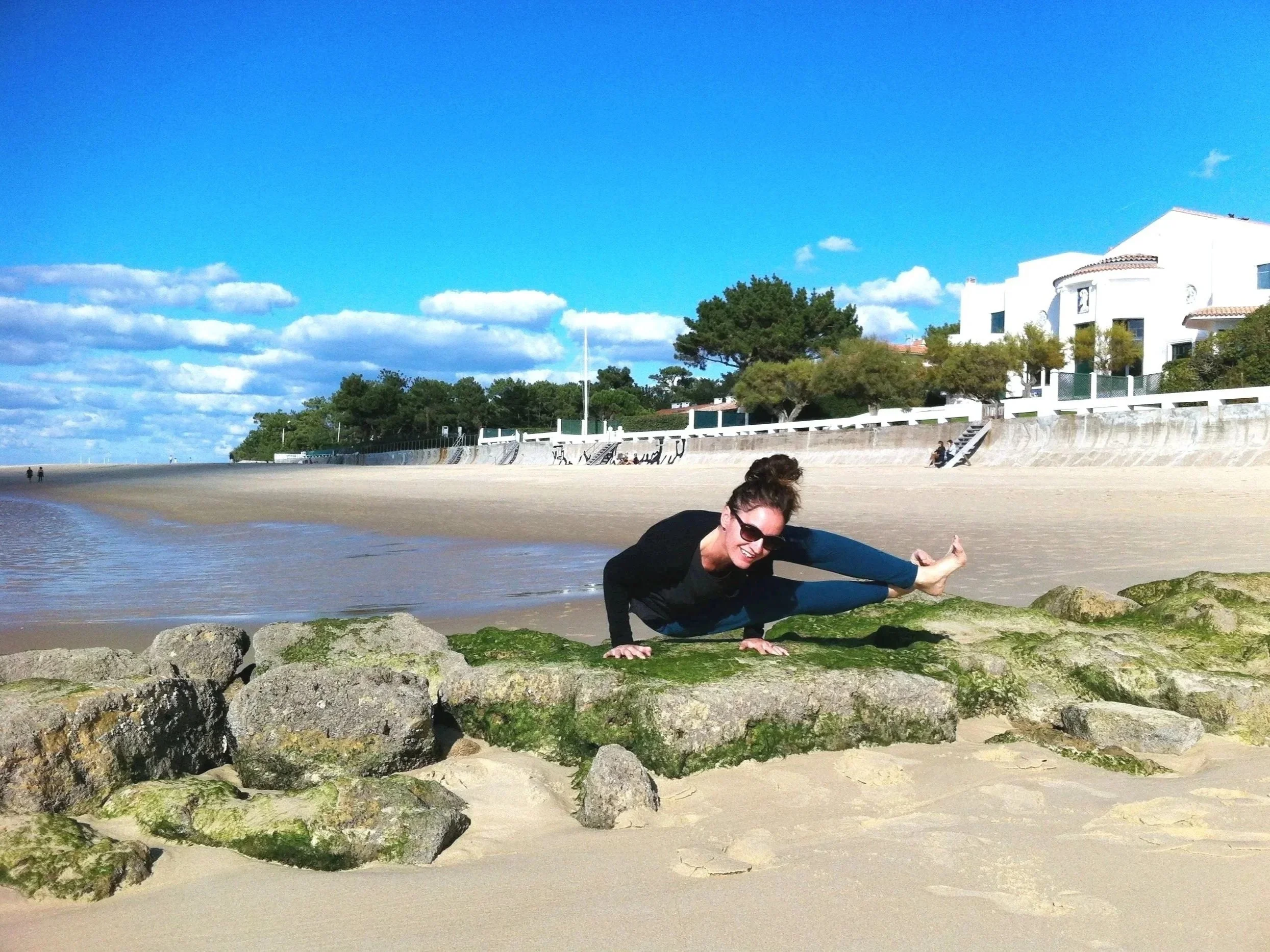 Ludivine en position de yoga sur un rocher au bord de la plage à Arcachon, souriante, portant des lunettes de soleil, avec une ville en arrière-plan et un ciel bleu avec quelques nuages.