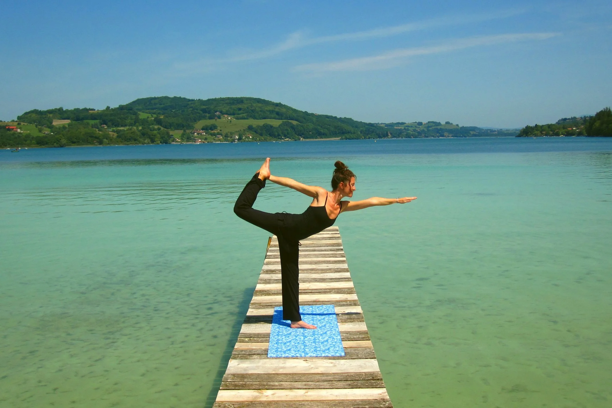 Une femme pratique le yoga sur une jetée en bois au bord d'un lac avec une vue sur une montagne verte et un ciel bleu.