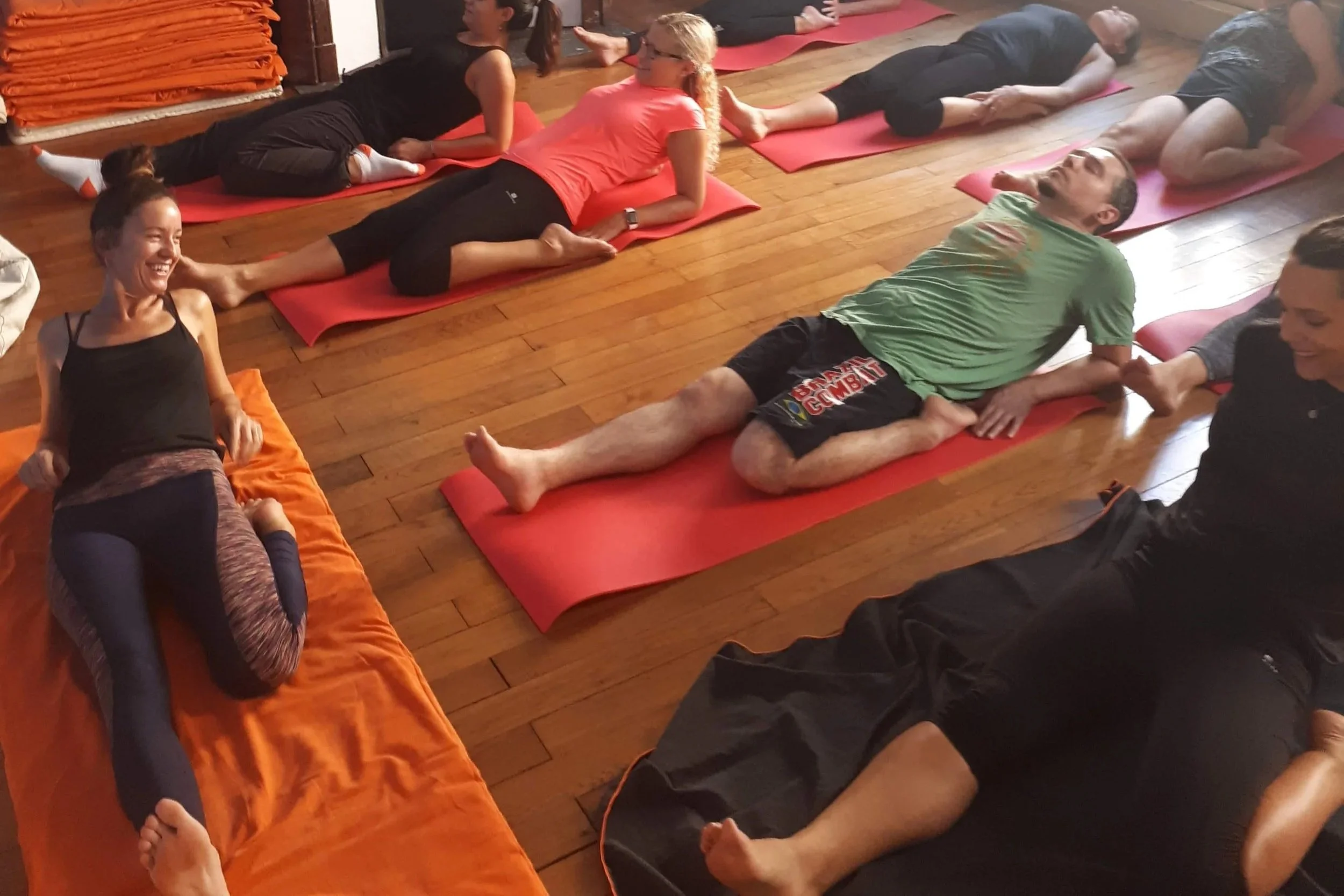 Groupe de personnes participant à une séance de yoga ou de stretching dans une salle en bois, certaines sont allongées et d'autres sont assises ou en position de relaxation.