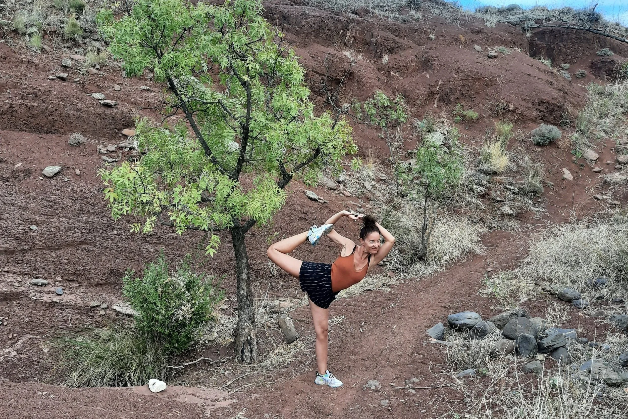 Une femme en tenue sportive effectue une posture d'étirement dans un paysage désertique avec quelques petits arbres et des rochers.