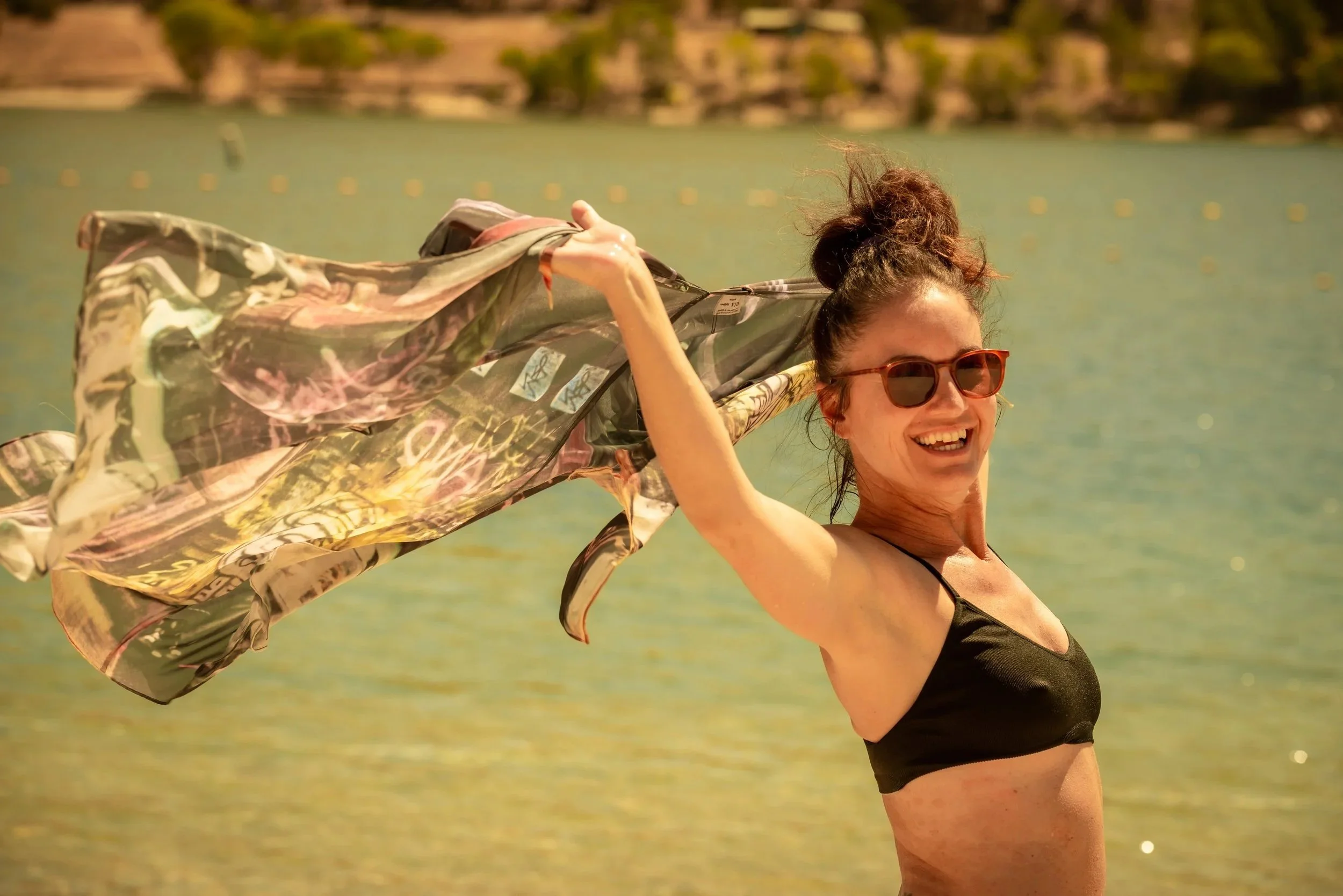Femme souriante portant des lunettes de soleil, portant un haut noir, se tenant sur la plage près de l'eau, avec un vêtement coloré qu'elle agite dans l'air.