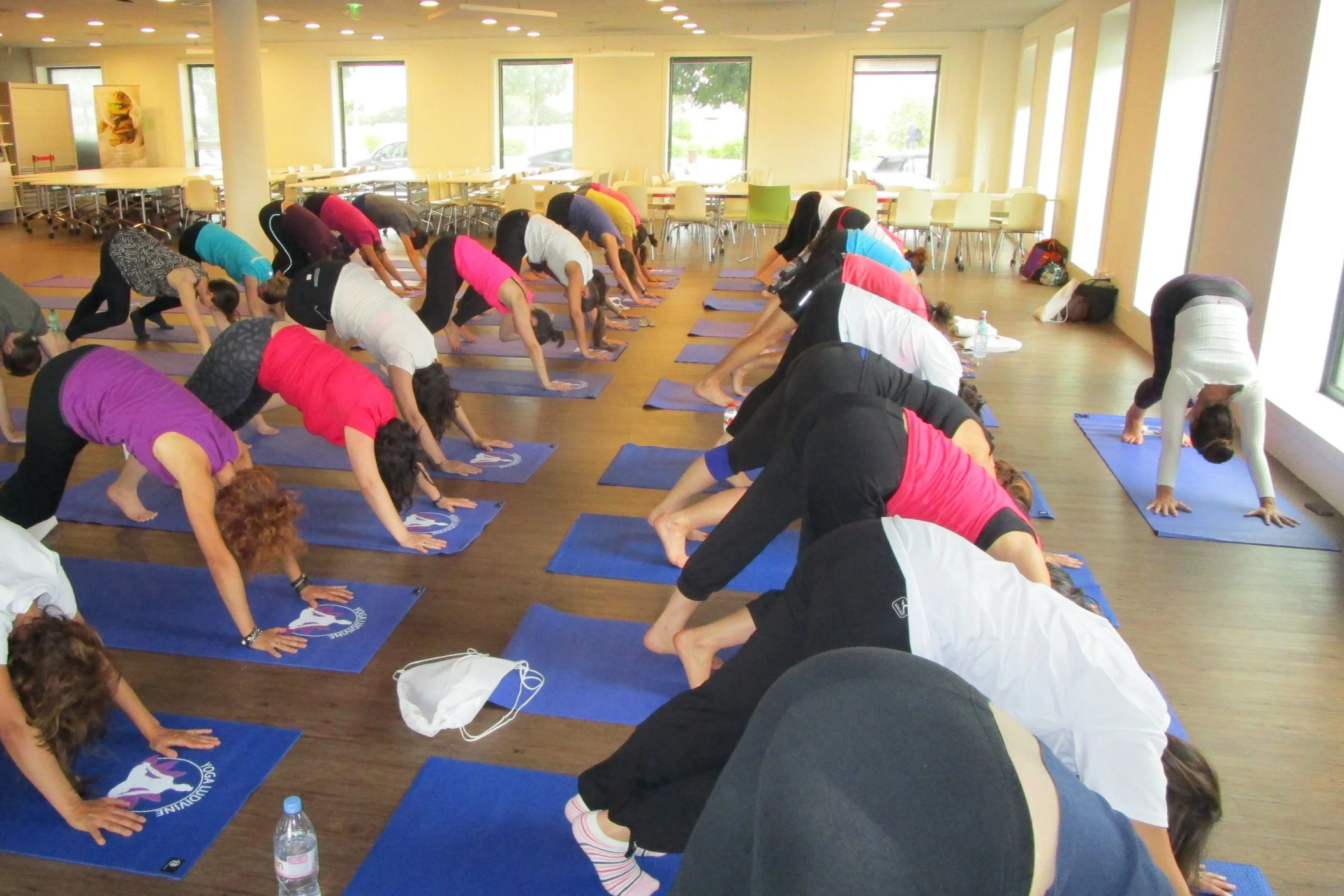 Groupe de personnes faisant du yoga dans une salle de sport lumineuse, en position de chien tête en bas, sur des tapis bleus.