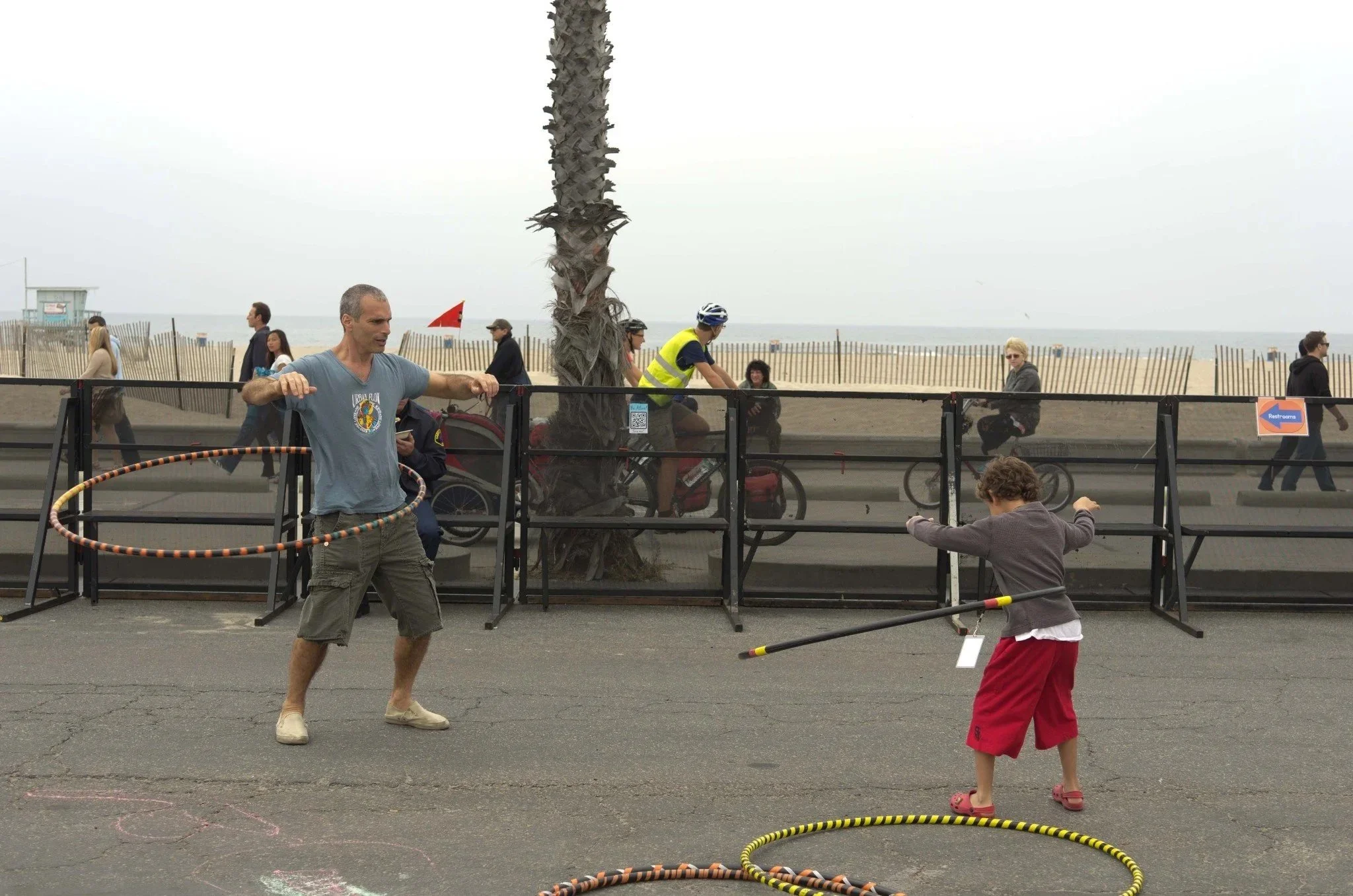 Un homme et un enfant jouent avec des hula hoops sur un trottoir face à la plage, avec des piétons et des cyclistes en arrière-plan.