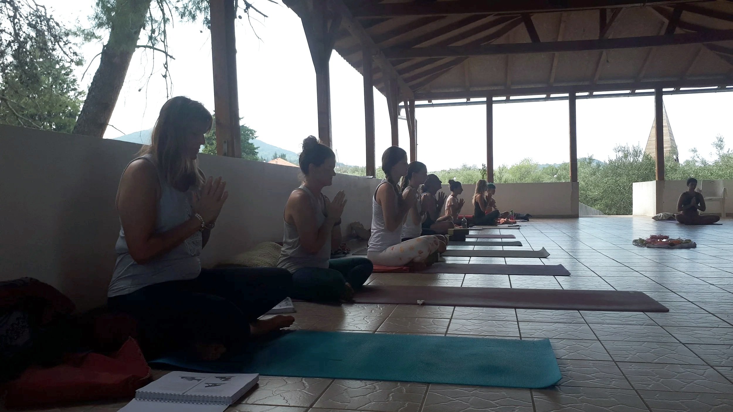 Groupe de personnes pratiquant la méditation en position assise dans une salle ouverte avec vue sur la nature.