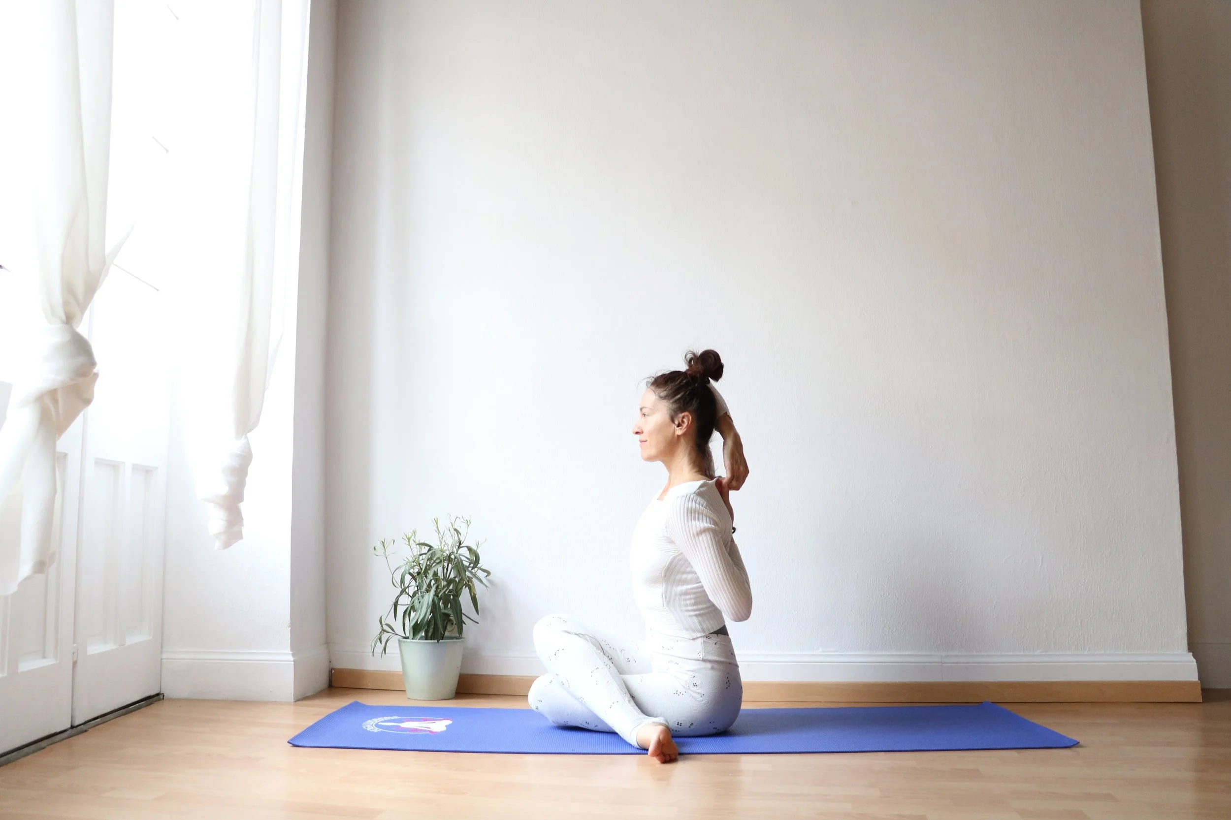 Femme pratiquant la relaxation ou la méditation assise sur un tapis de yoga bleu dans une pièce lumineuse et minimaliste.