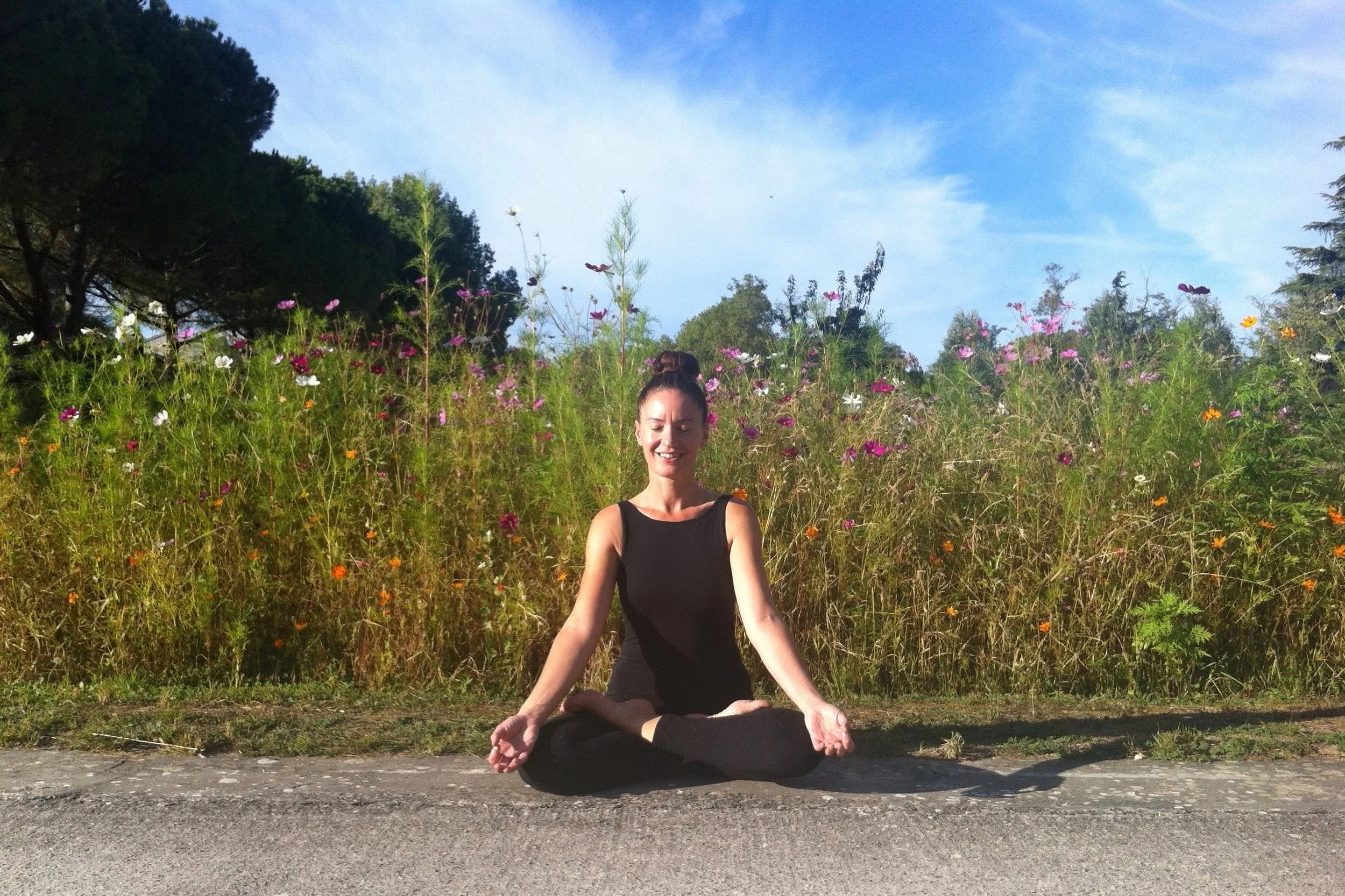 Une femme en habit noir pratiquant la méditation en position du lotus sur le sol en plein air, avec un champ de fleurs colorées et des arbres en arrière-plan, sous un ciel bleu avec quelques nuages.