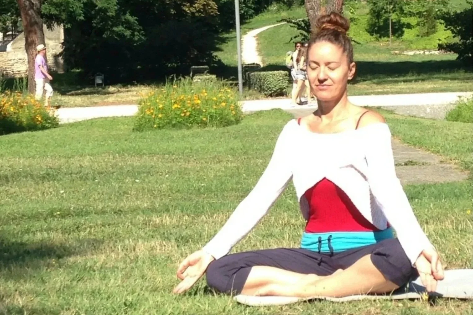 Femme méditant en position de lotus dans un parc ensoleillé, arbres et fleurs en arrière-plan.