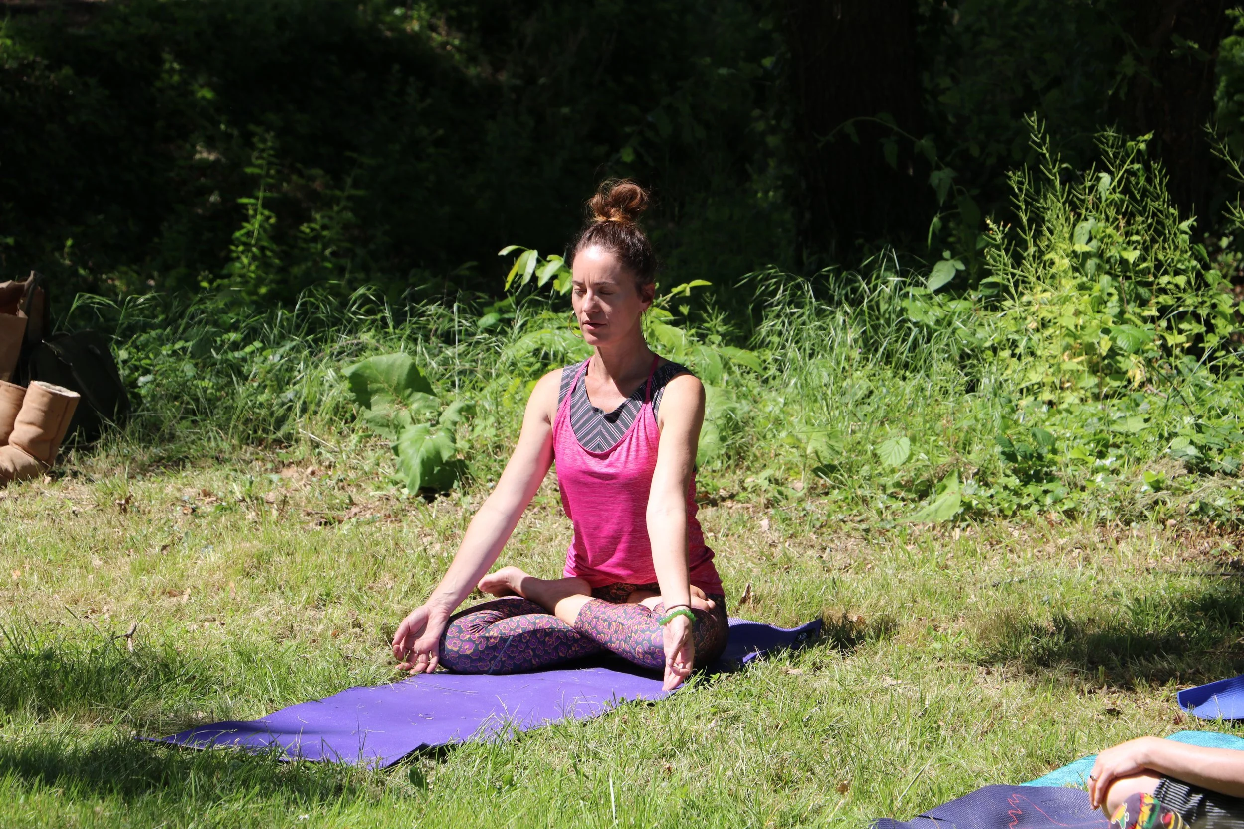 Une femme en train de faire du yoga en plein air, assise en position de méditation sur un tapis violet, dans un espace herbeux avec des arbres et des plantes derrière elle.