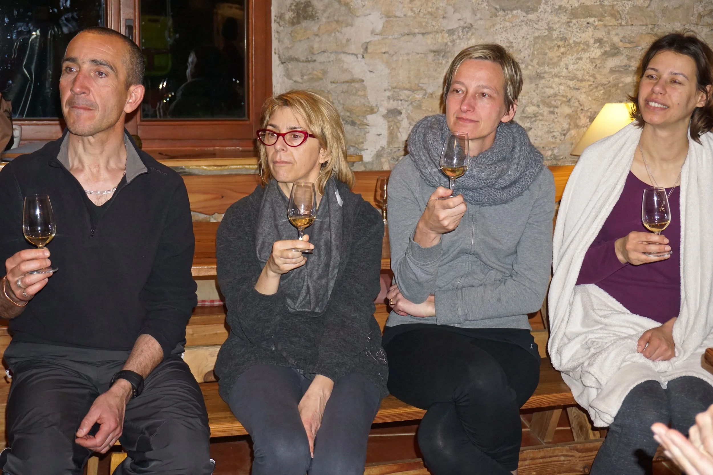 Four people are sitting on a wooden bench holding glasses of wine during a gathering in a cozy indoor setting. The group includes three women and one man, all appearing to enjoy a social moment.