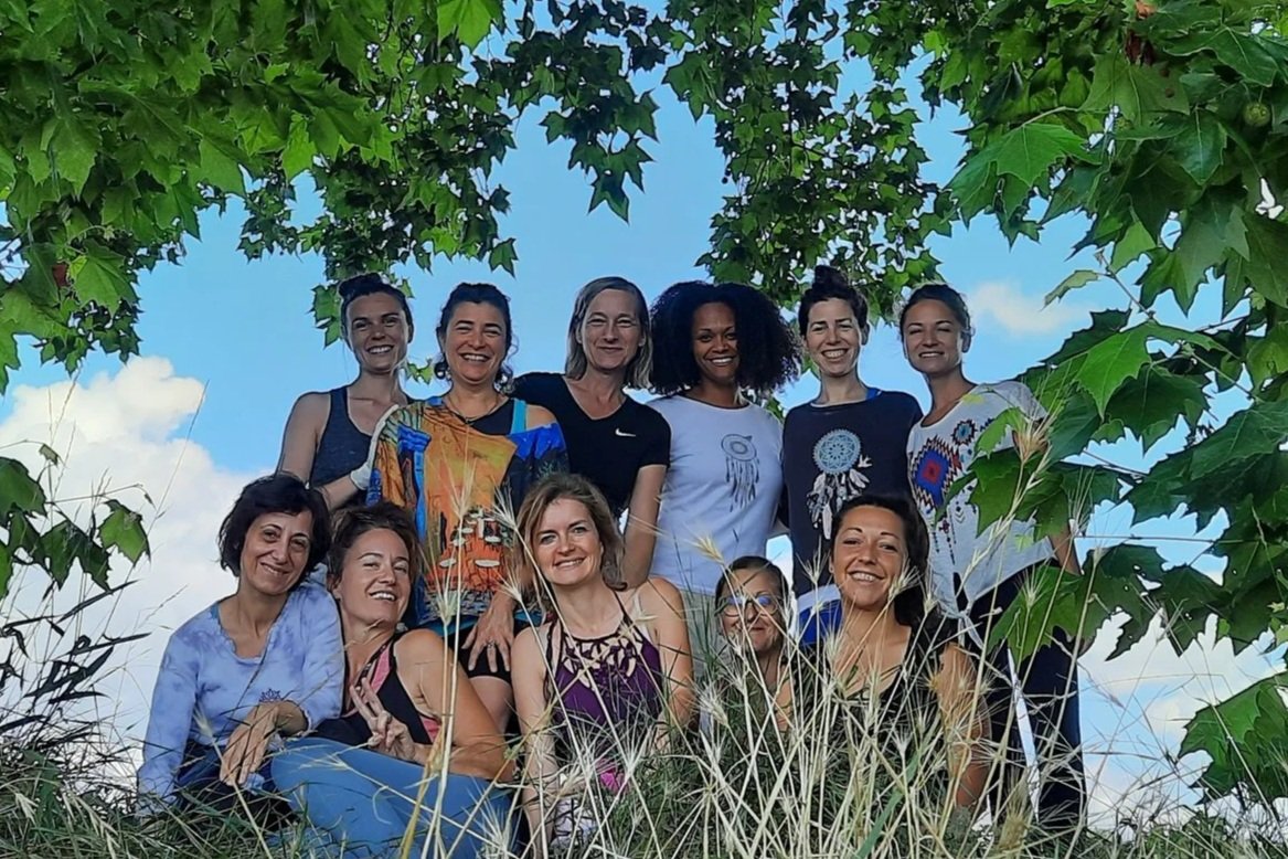 Un groupe de 12 femmes souriantes posant sous un arbre en plein air, avec un ciel bleu et quelques nuages en arrière-plan.