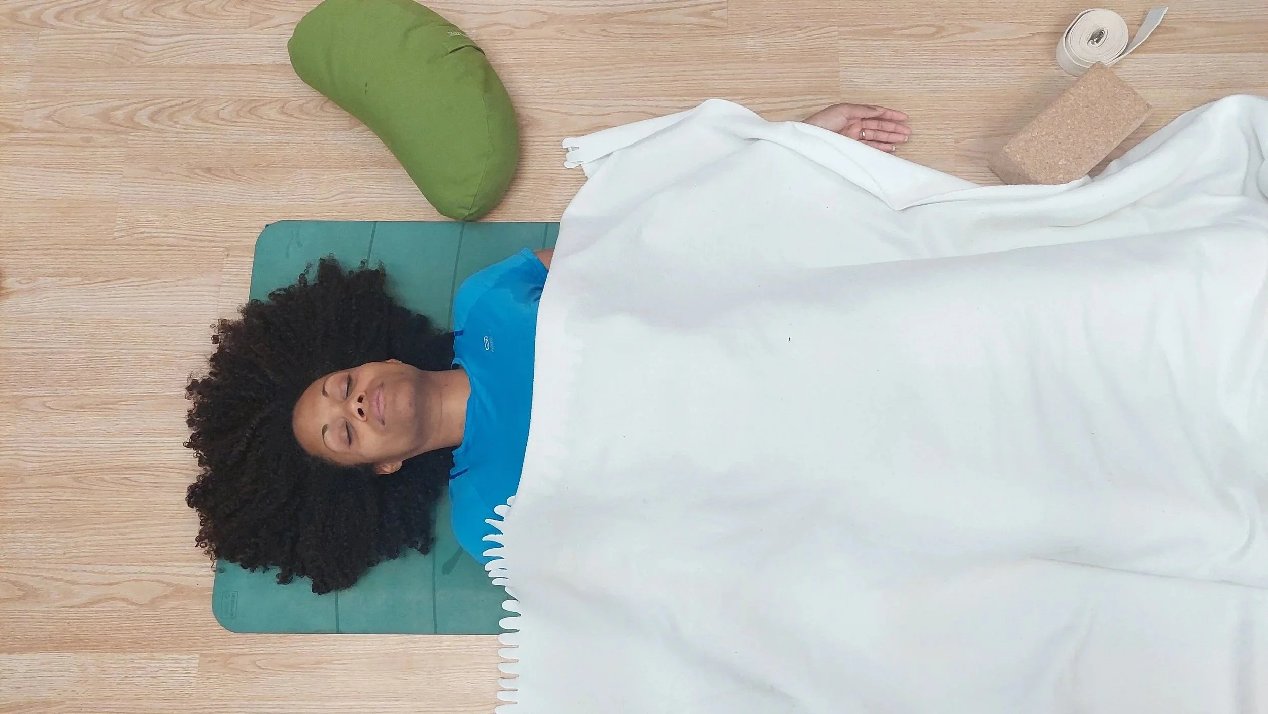 Élève allongée sur un tapis de yoga en Savasana posture de relaxation, yeux fermés, avec un coussin vert à côté, recouverte d'une couverture blanche, dans une pièce avec un sol en bois.