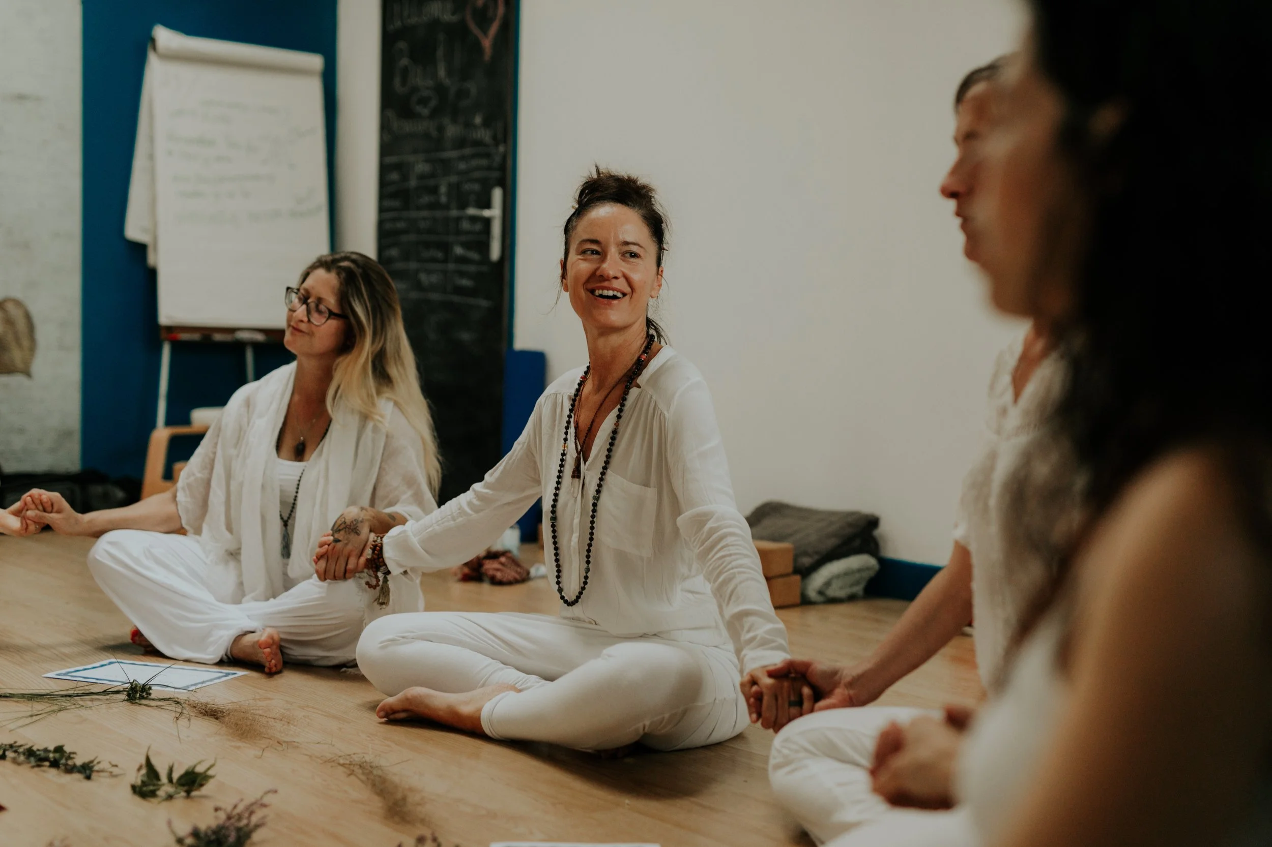 Groupe de Yoginis assises en cercle sur le sol, participant à une séance de méditation et de yoga, dans une pièce calme avec un tableau blanc au mur. Durant notre formation de professeur de yoga.