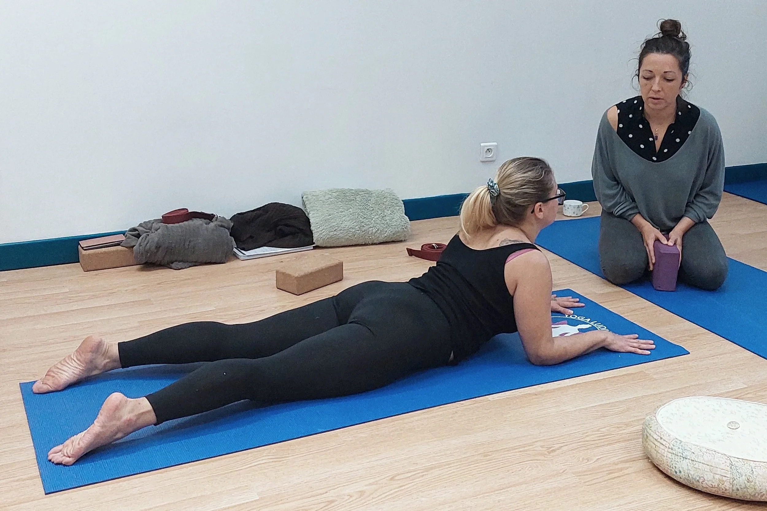 Une femme en position de Cobra pendant une séance de yoga, avec une autre femme en face aidant avec un bloc.