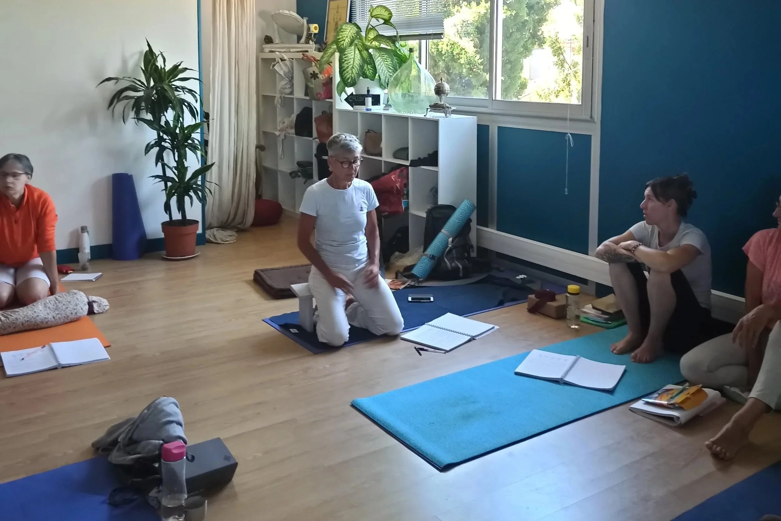 Groupe d'élèves assises sur des tapis de yoga dans une salle lumineuse avec des plantes et des étagères enseignant pendant la formation de professeur de yoga.