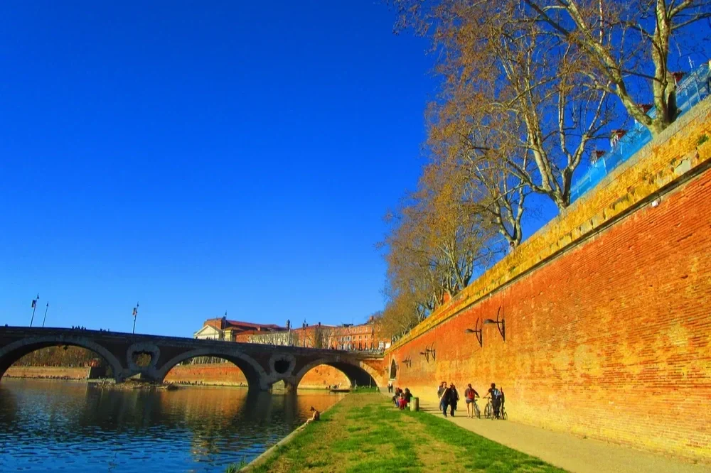 Promenade au bord de la rivière avec des personnes, des arbres sans feuilles et un vieux mur de briques rouges sous un ciel bleu clair.