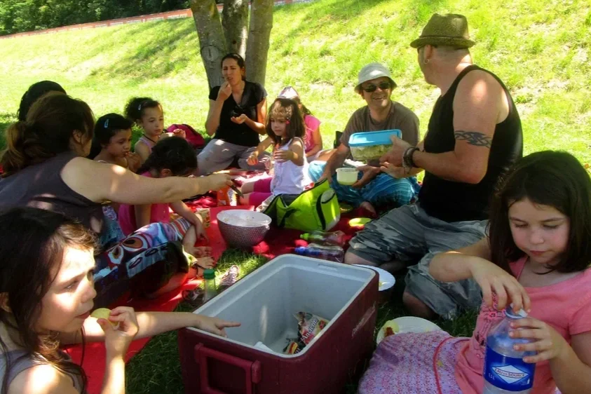 Groupe de personnes, adultes et enfants, en pique-nique sous un arbre, avec de la nourriture, des boissons et un sac à dos, en plein air sous le soleil.