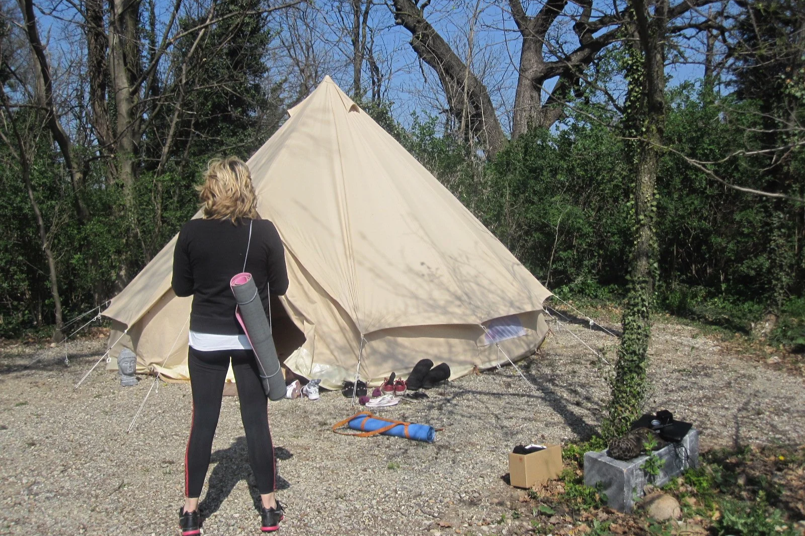 Une femme vue de dos, positionnée devant une grande tente beige dans un environnement boisé. Elle porte des vêtements de sport noirs et une corde à saillie à l'épaule. Autour de la tente, il y a des chaussures, un tapis de yoga ou de camping, une boî