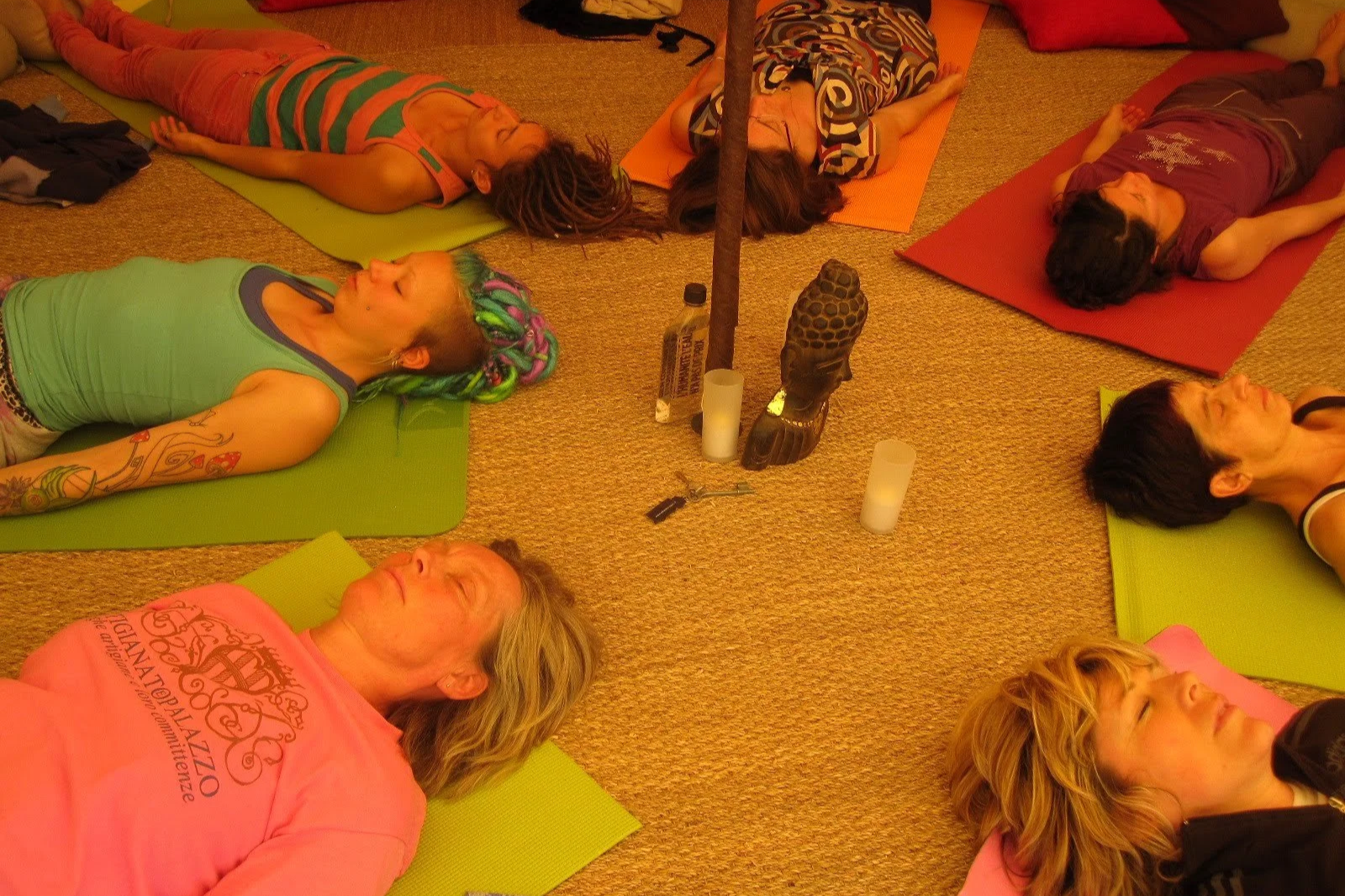 Groupe de personnes en yoga ou méditation allongées sur des tapis colorés, dans une salle, entourées de bougies, avec des chaussures et des objets au centre.