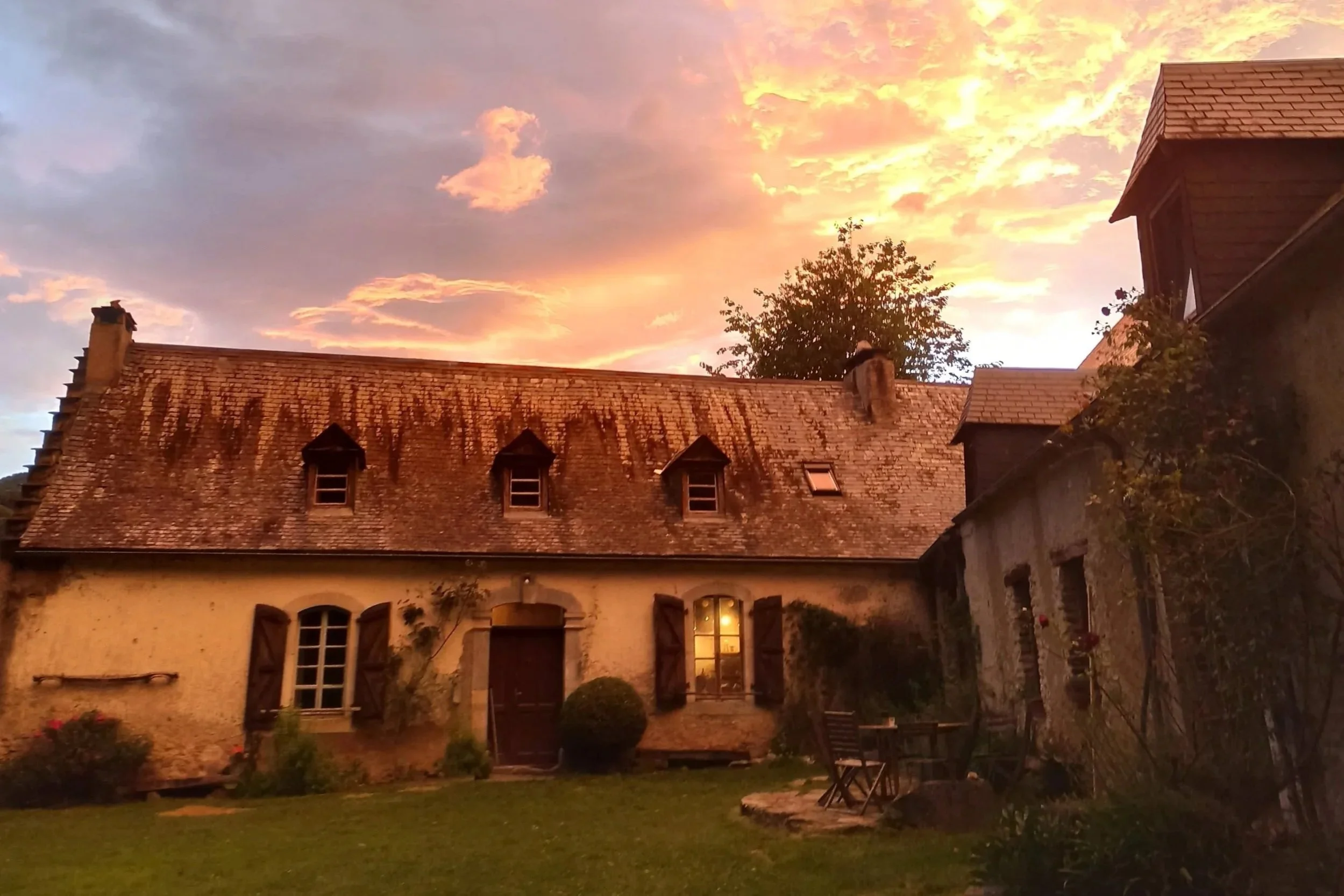 Une vieille maison en pierre avec un toit en tuiles, des fenêtres à volets en bois, un jardin devant avec des meubles en bois, sous un ciel coucher de soleil aux couleurs chaudes.